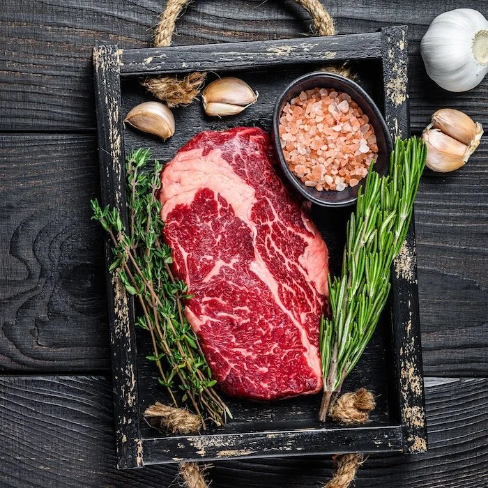 Raw ribeye steak with fresh rosemary, thyme, garlic, and pink salt in a rustic wooden tray — ranch-direct beef from Dry Creek Pastures.