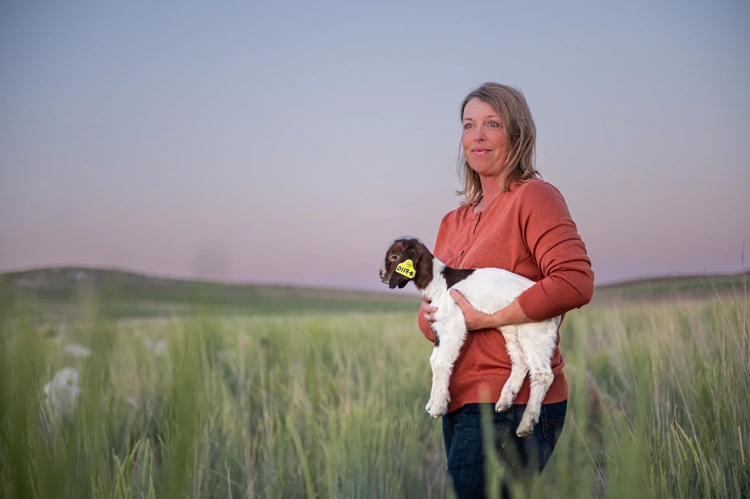 Millie Bradshaw of Dry Creek Pastures holding a Boer kid in a green pasture at dusk — Woodruff, Utah.