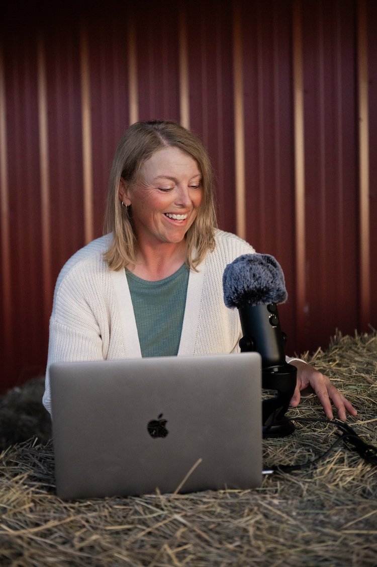 Millie Bradshaw recording the Goat Wise podcast with laptop and microphone on hay bales in a barn at Dry Creek Pastures