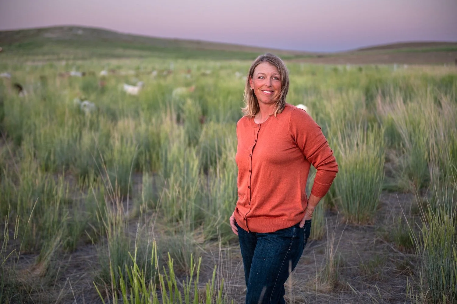 Millie Bradshaw smiling in tall grass meadow with meat goat herd grazing in background at Dry Creek Pastures at dusk