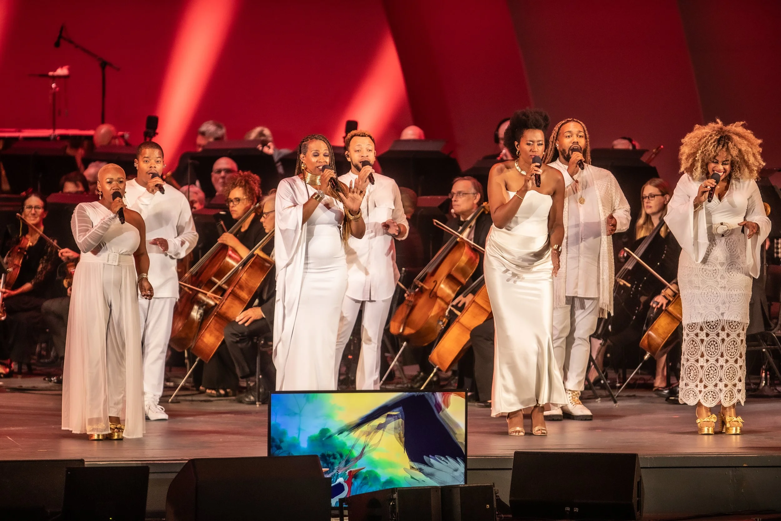 A group of six singers performing on stage with orchestra behind them, all dressed in white, with red lighting and a colorful display on a monitor in front.