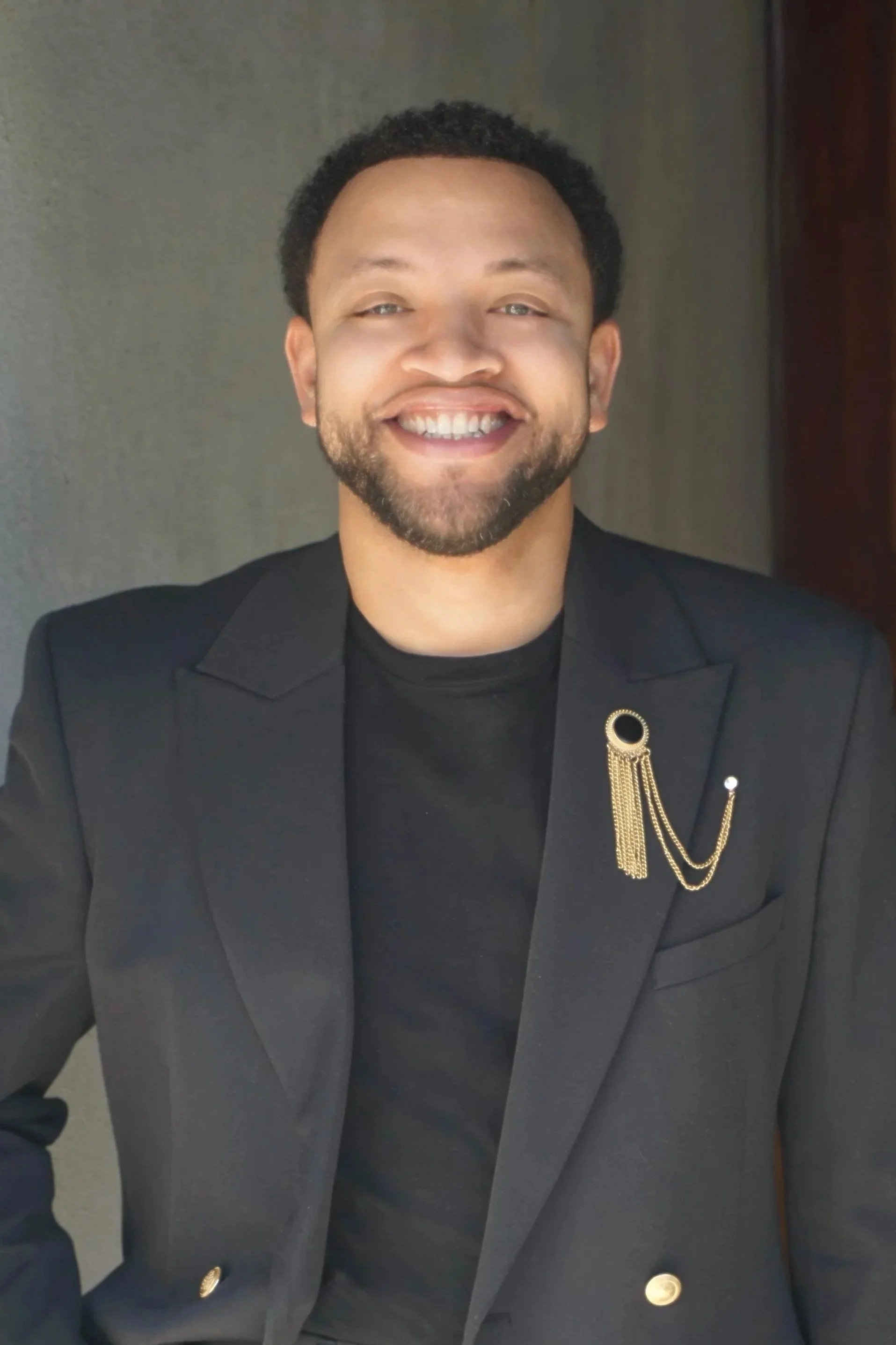 A smiling man with a beard and mustache wearing a black blazer with gold chain and brooch, sitting indoors.