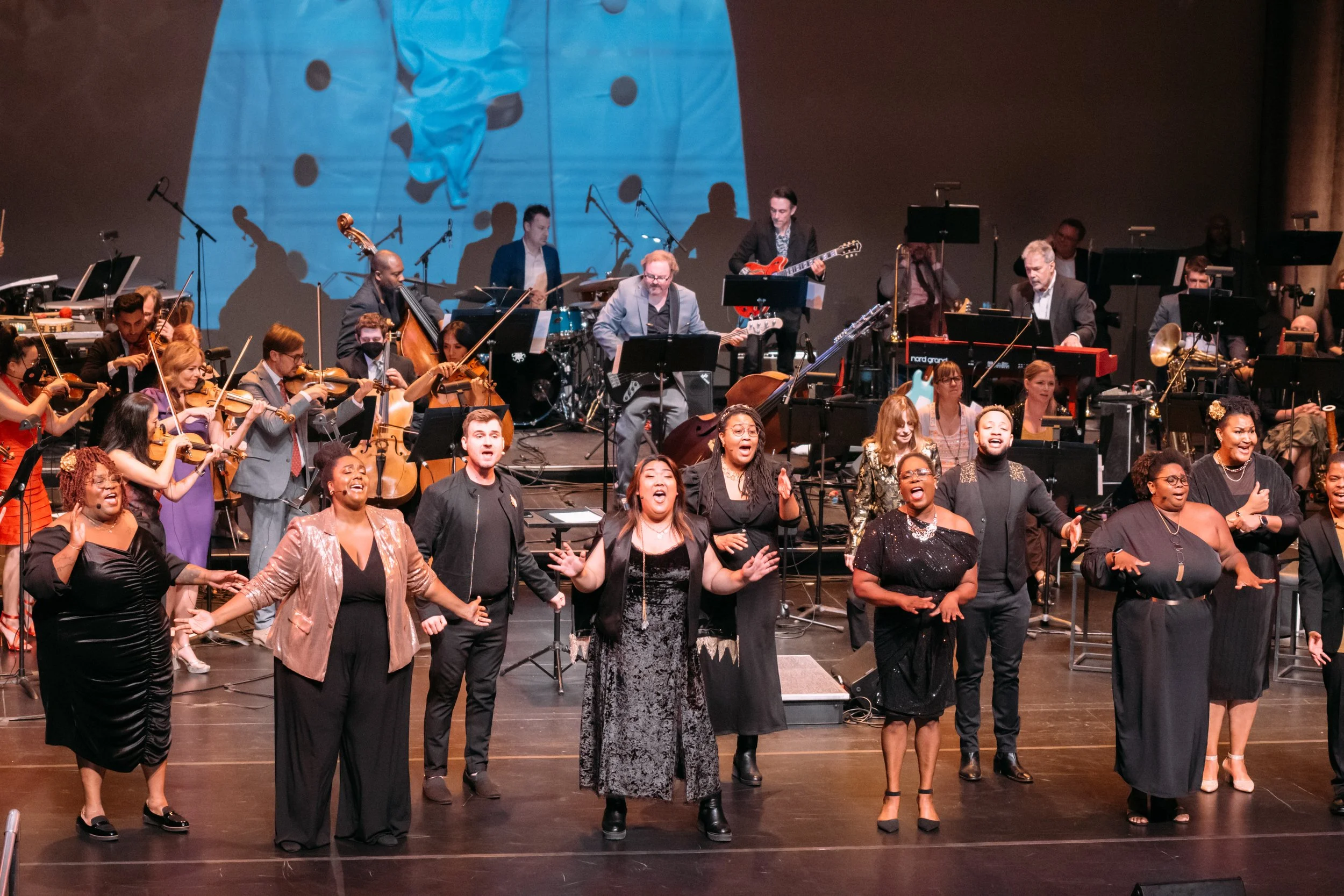 A diverse group of singers and musicians on stage performing with orchestral musicians in the background.