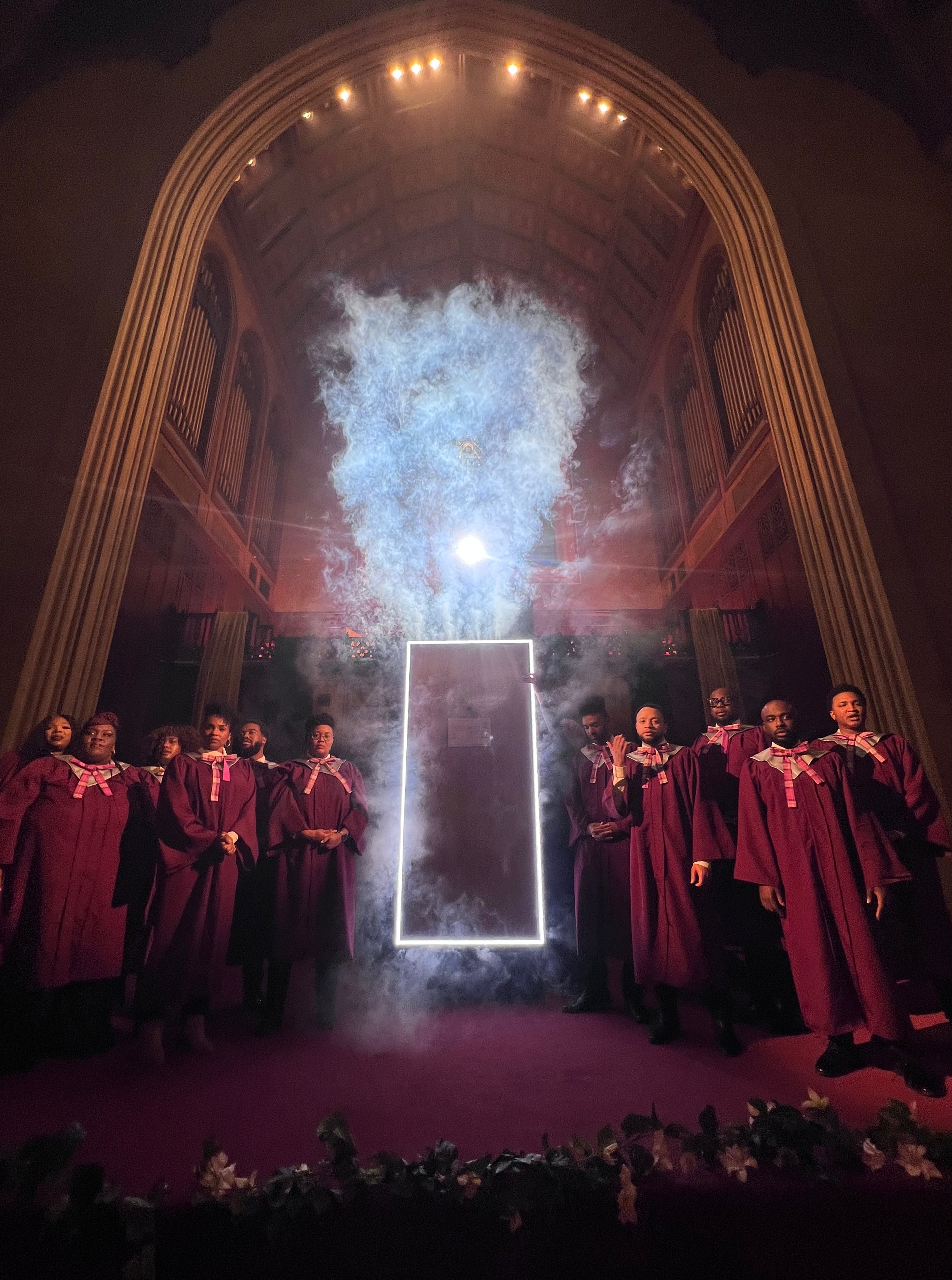 Group of singers in maroon gowns standing on a stage in a church with a high, arched ceiling, smoke and bright light behind a rectangular frame.
