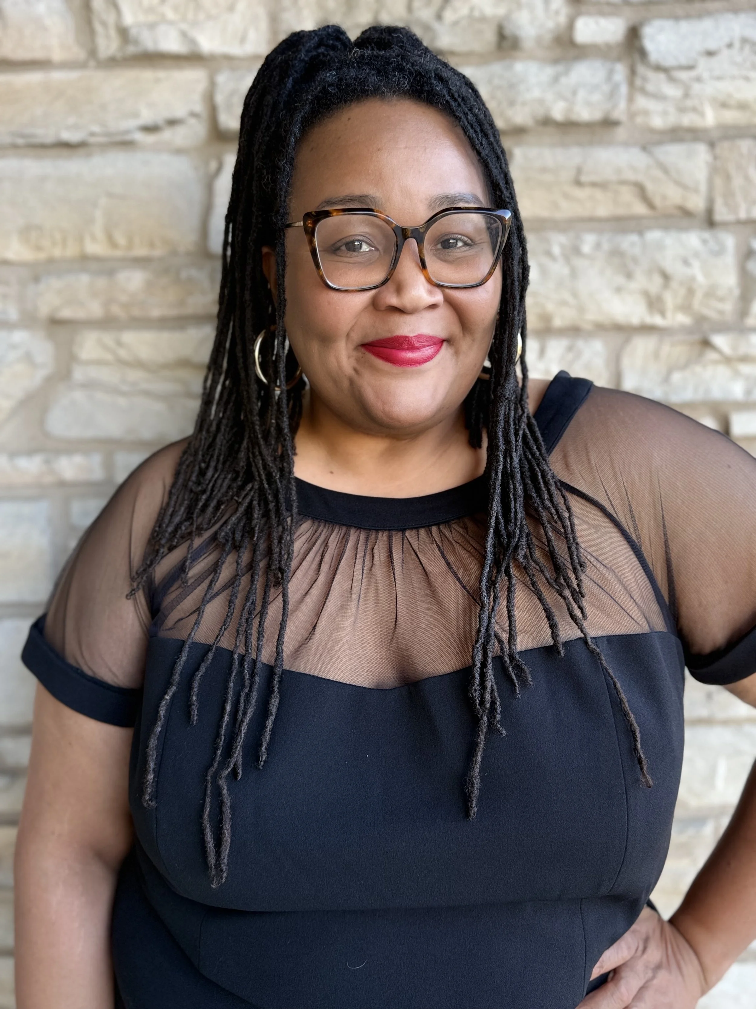 A woman with glasses and dreadlocks wearing a black dress with sheer brown top, standing in front of a brick wall, smiling.