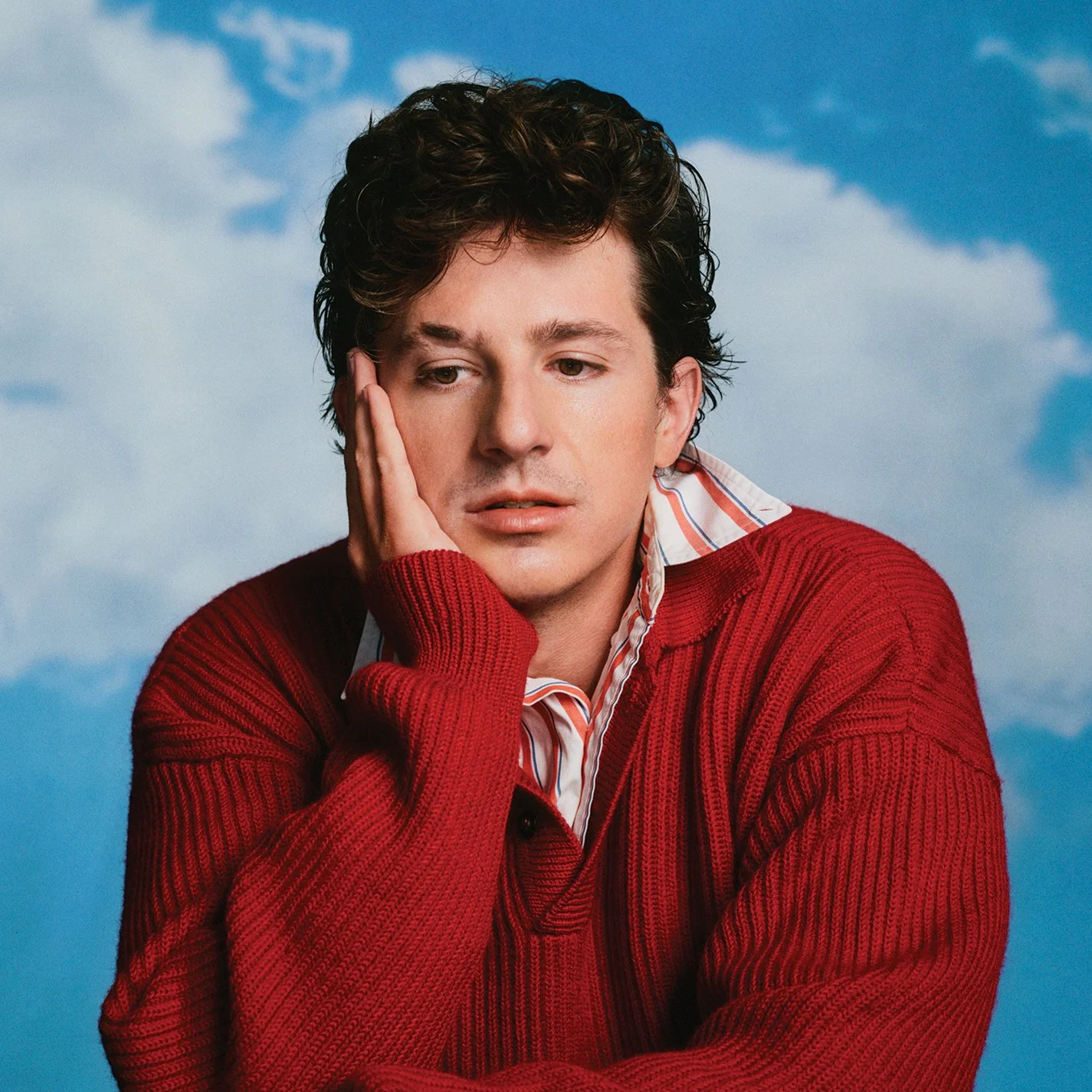 A young man with curly brown hair, wearing a red sweater, holding his face with one hand, looking pensive against a cloudy sky background.