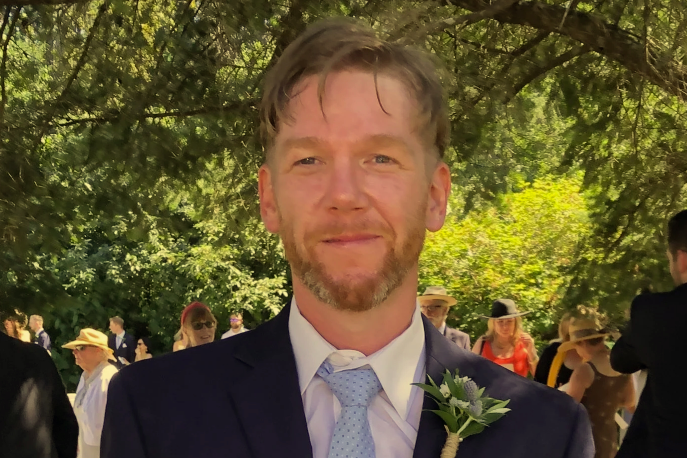 A man with light brown hair and a beard, dressed in a dark suit, white shirt, and light blue tie, standing outdoors under a leafy canopy at a social event, with several people dressed formally in the background.