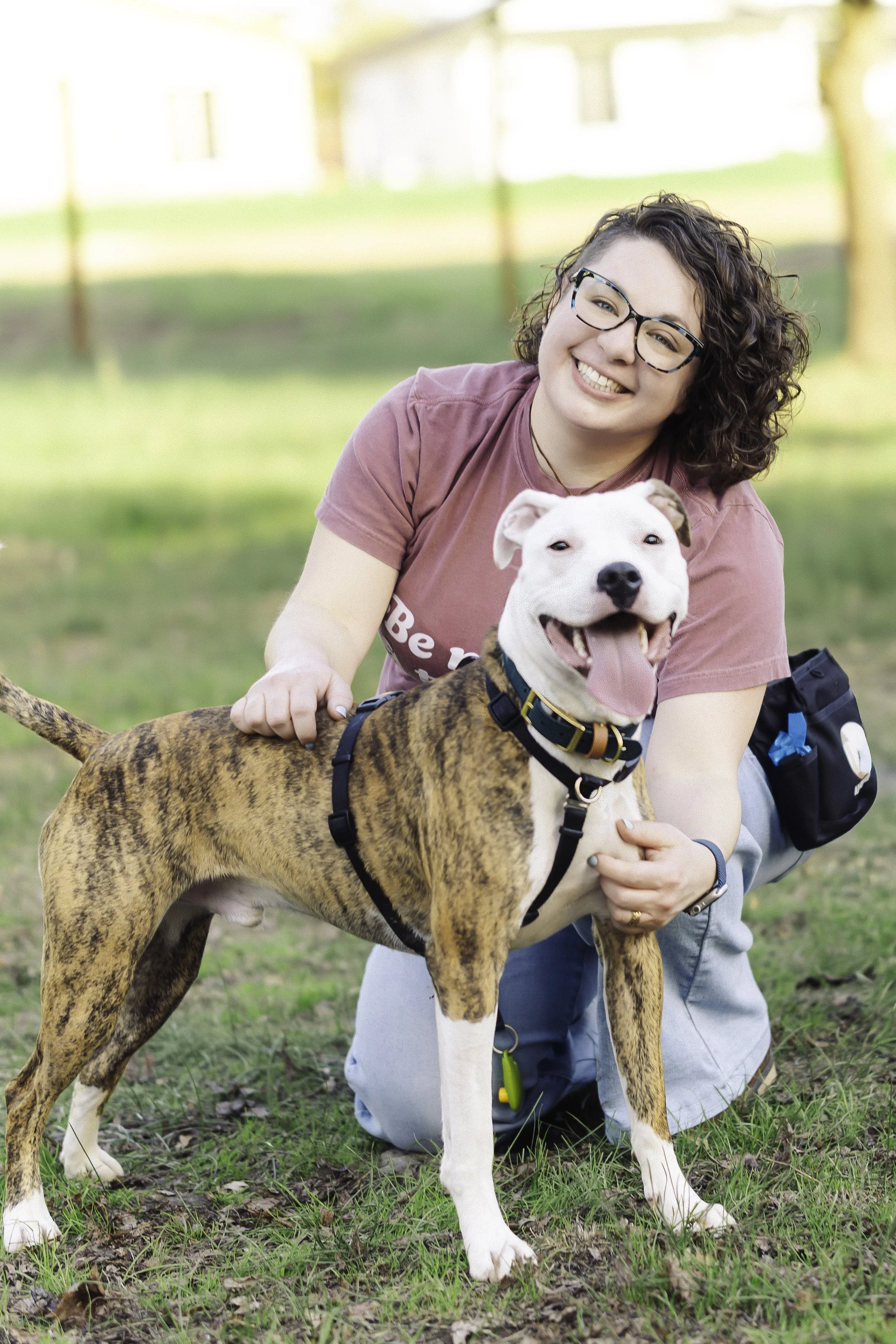 A woman with curly hair and glasses smiling, kneeling outside on grass with her brindle and white dog, both looking happy and playful.