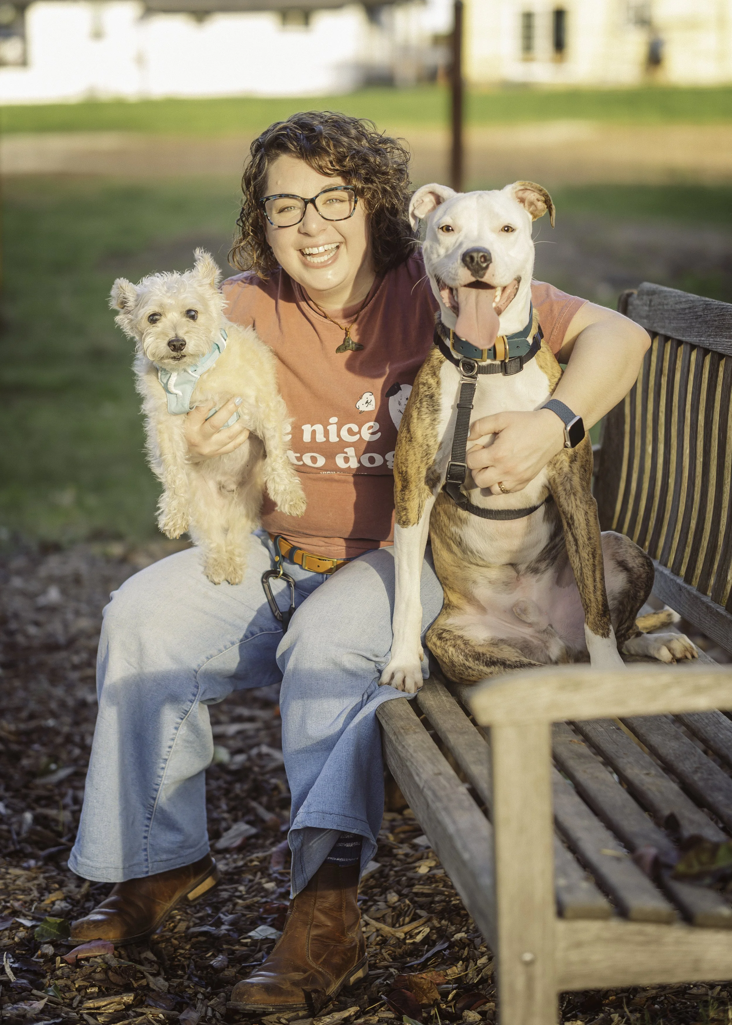 A woman sitting on a park bench holding two dogs, one small white dog wearing a light blue bandana, and a large brindle and white dog with its tongue out. The woman is smiling and wearing glasses, a maroon T-shirt, jeans, and brown boots.