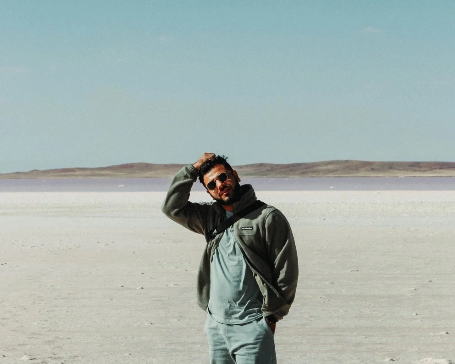 A man standing in a vast, open, salt flat landscape with dry hills in the background, wearing sunglasses, a gray jacket, and light-colored pants, with one hand on his head.