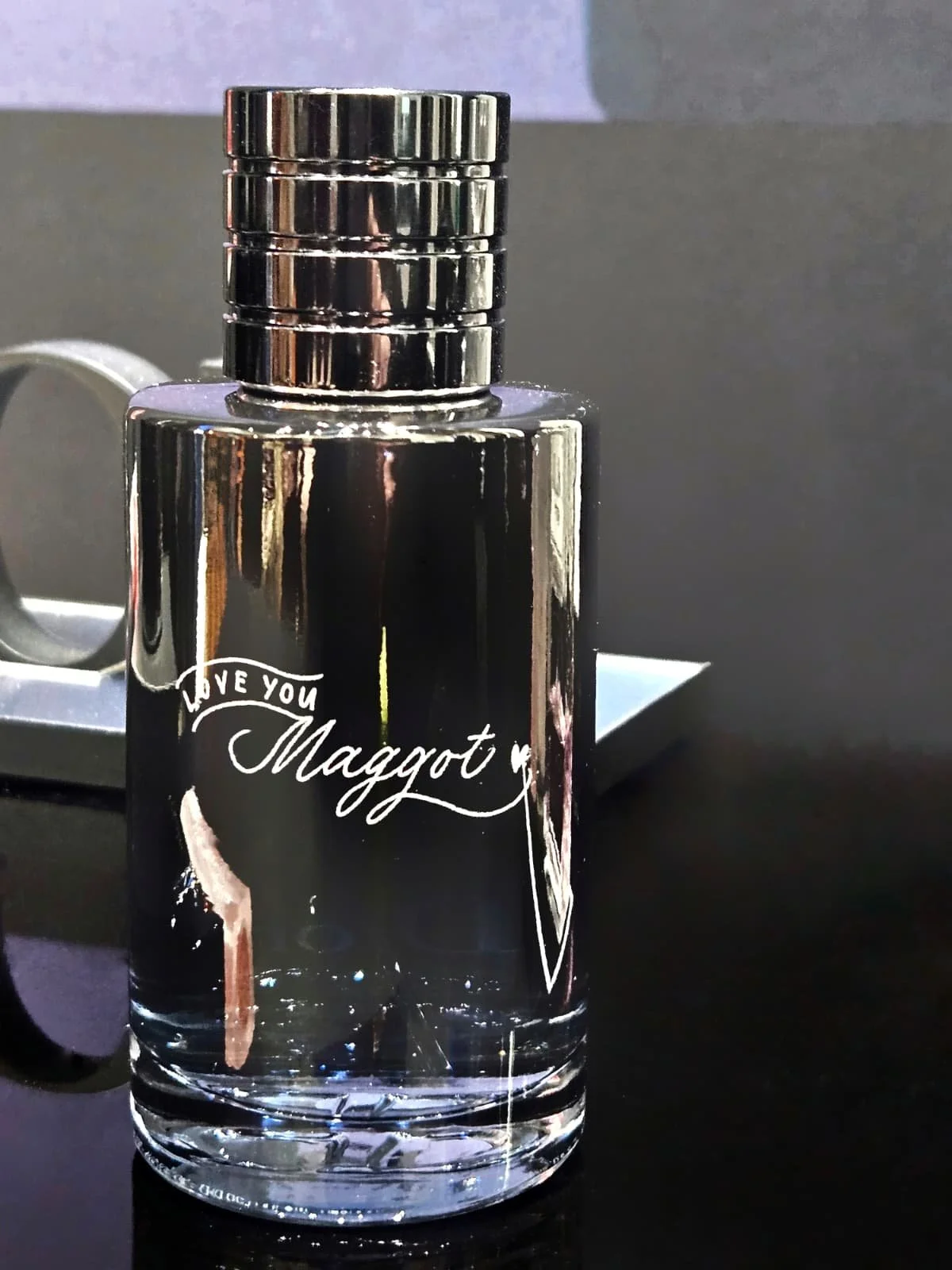A small glass perfume bottle with black label, silver cap, and decorative writing that reads 'Love You Maggot' with a small heart.