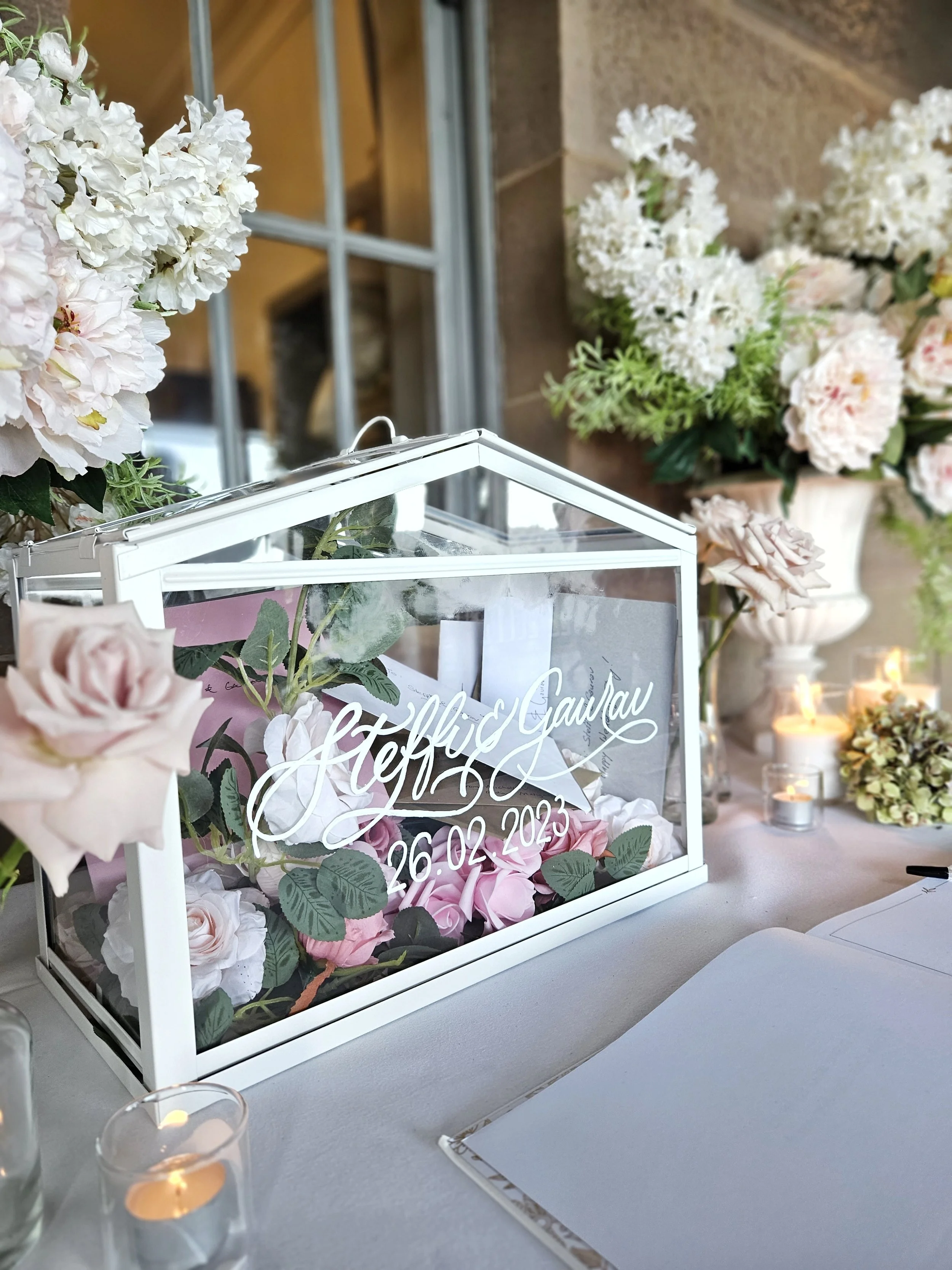 Glass display case with white calligraphy that says 'Steffi & Gawhar, 26.02.2023', surrounded by pink and white roses and green leaves, on a table decorated with candles and flowers, with a background of flowers and a window.