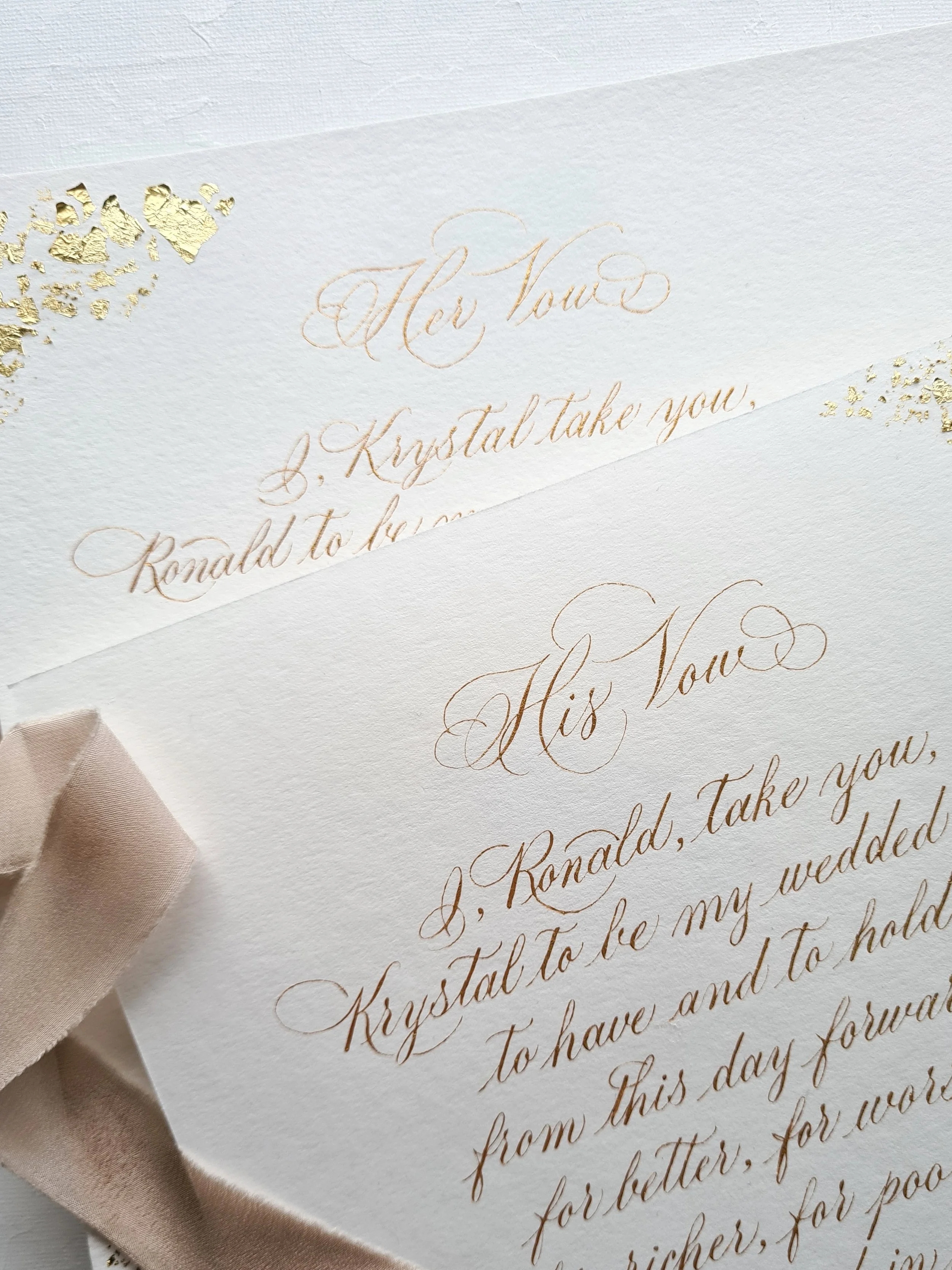 Close-up of wedding invitation cards with elegant gold calligraphy and beige ribbon, partially overlapped.