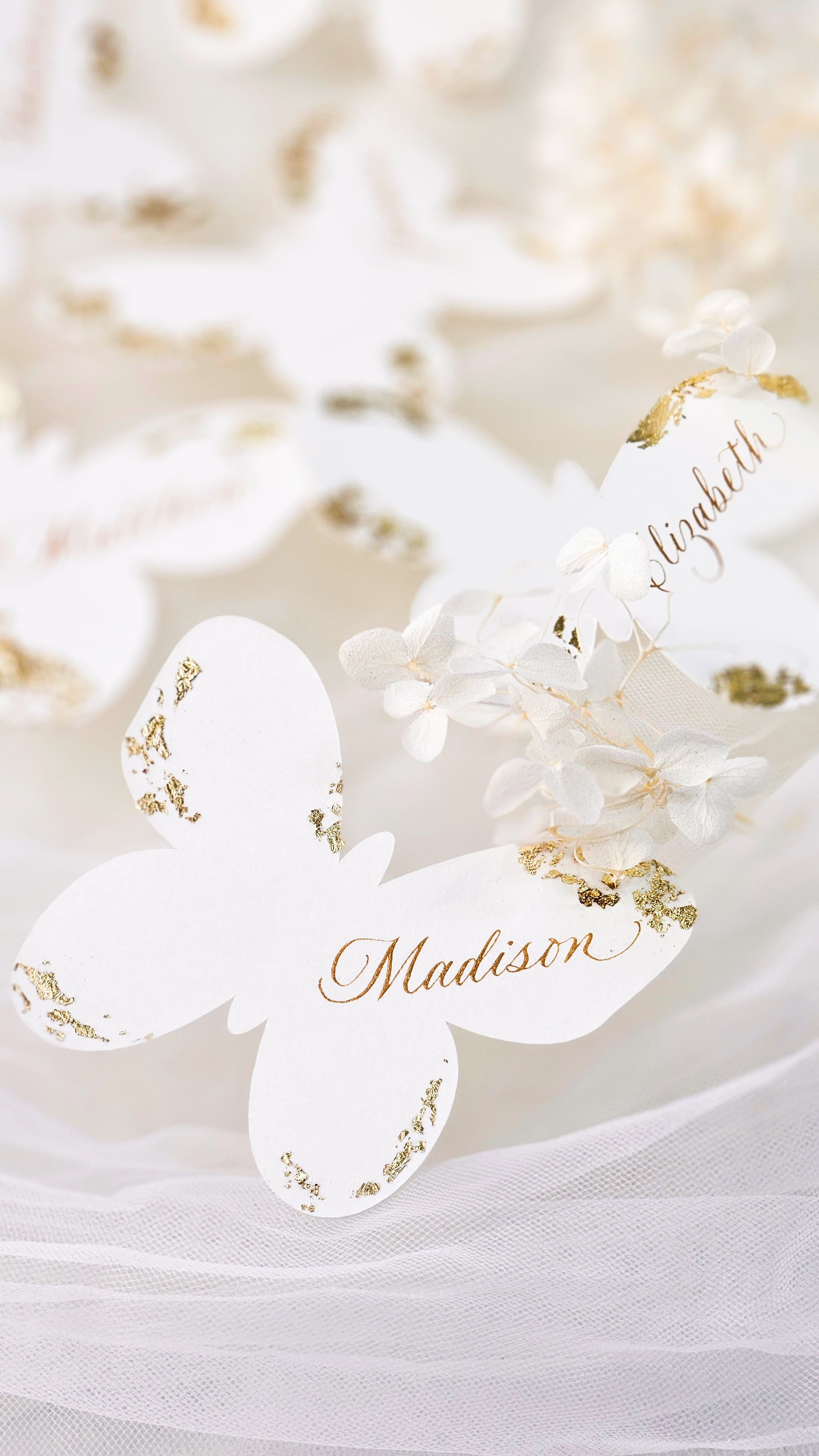 White custom butterfly placecards with 'Madison' calligraphy on it, surrounded by white flowers and gold accents, on a soft white fabric background.