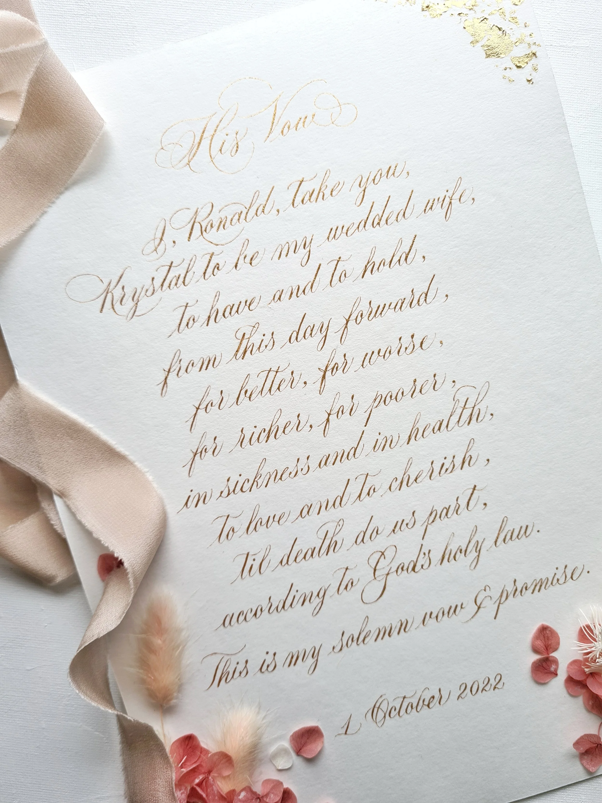 A wedding vow written in elegant calligraphy on white textured paper, decorated with pink flower petals, a soft pink ribbon, and small white and beige dried flowers.