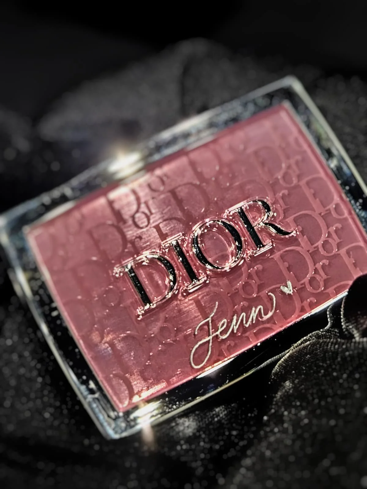 Close-up of a pink Dior eyeshadow palette with the name 'Jenna' written on it, placed on a black sparkly fabric.