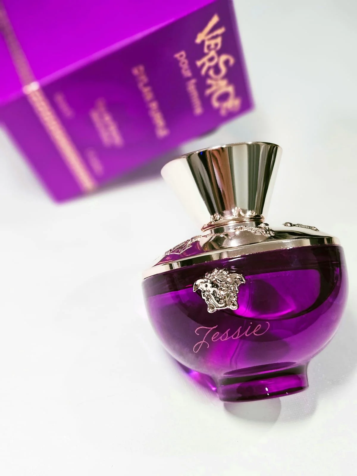 A purple perfume bottle with silver accents and the name 'Jessie' written on it, placed on a white surface with purple packaging blurred in the background.