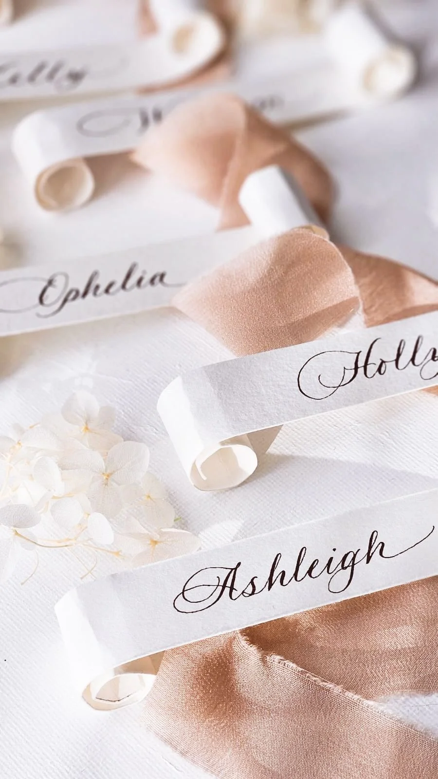 Close-up of custom name tags with elegant calligraphy, draped over a pink satin fabric and white textured surface, with white hydrangea flowers in the background.