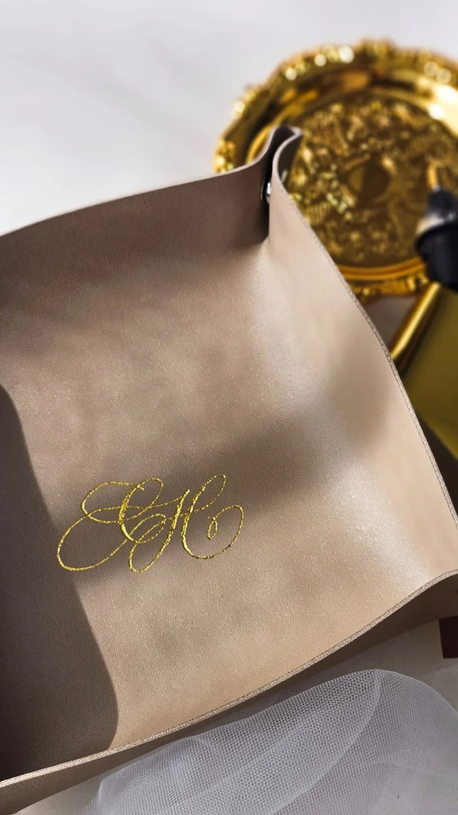 Close-up of beige fabric with gold embroidered initials 'HG', with a gold sequined accessory in the background.