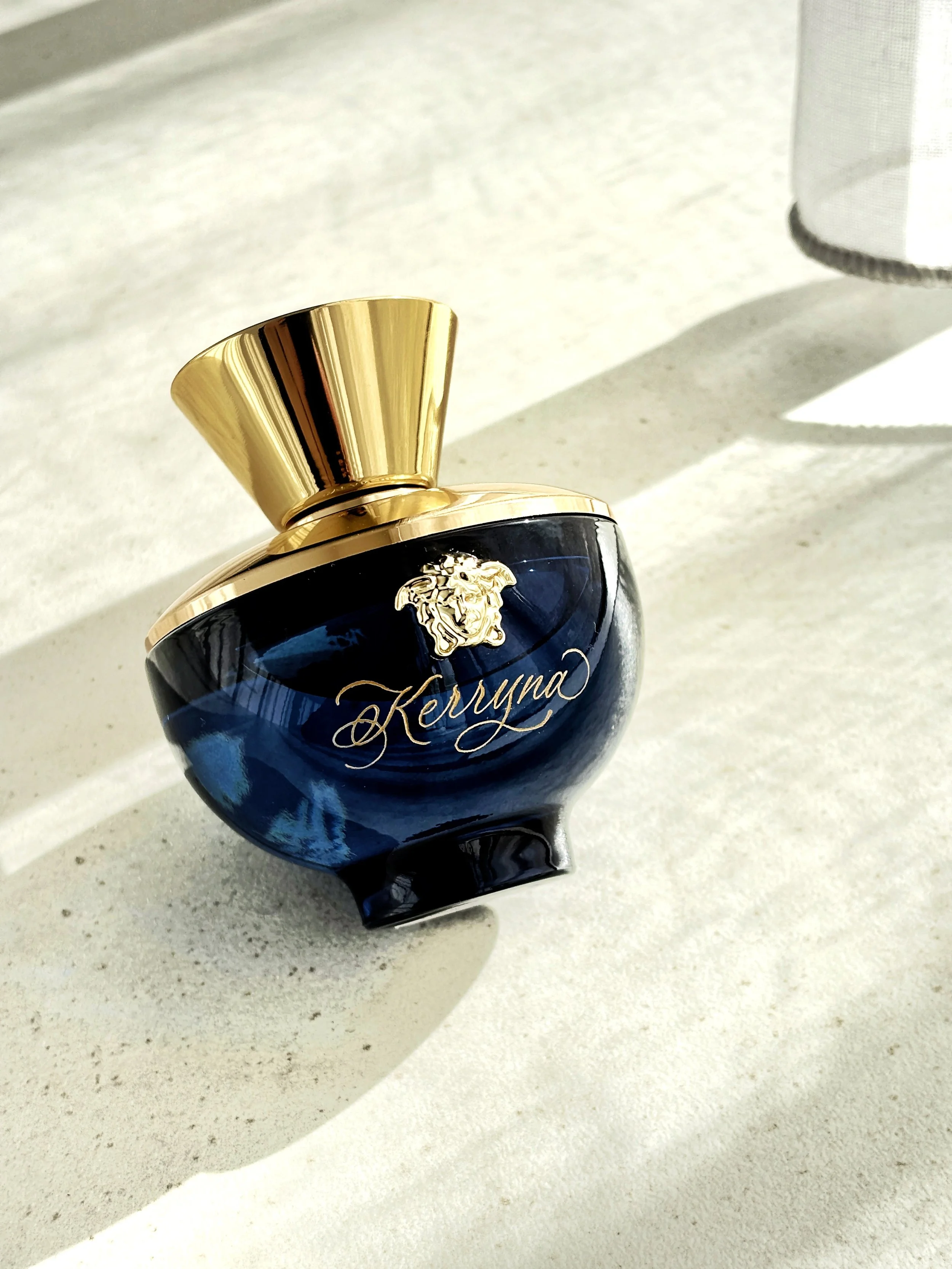 A luxurious perfume bottle with a navy blue body and gold accents, featuring the name 'Kreyana' and a lion head emblem, placed on a light-colored surface with sunlight and shadow.