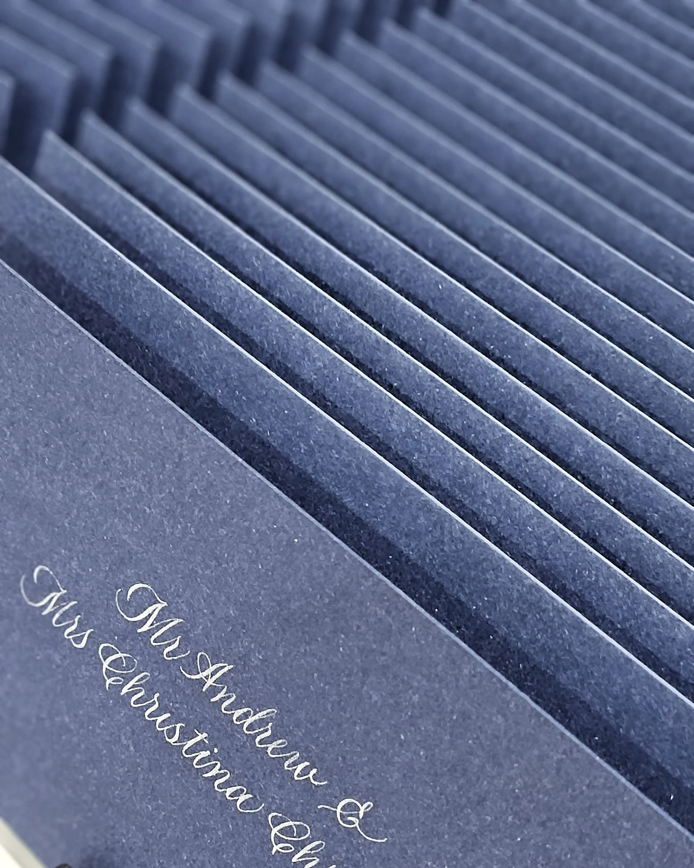 Close-up of navy blue wedding invitations with elegant white script, displaying the full names and titles of the couple, and featuring textured, ribbed paper.