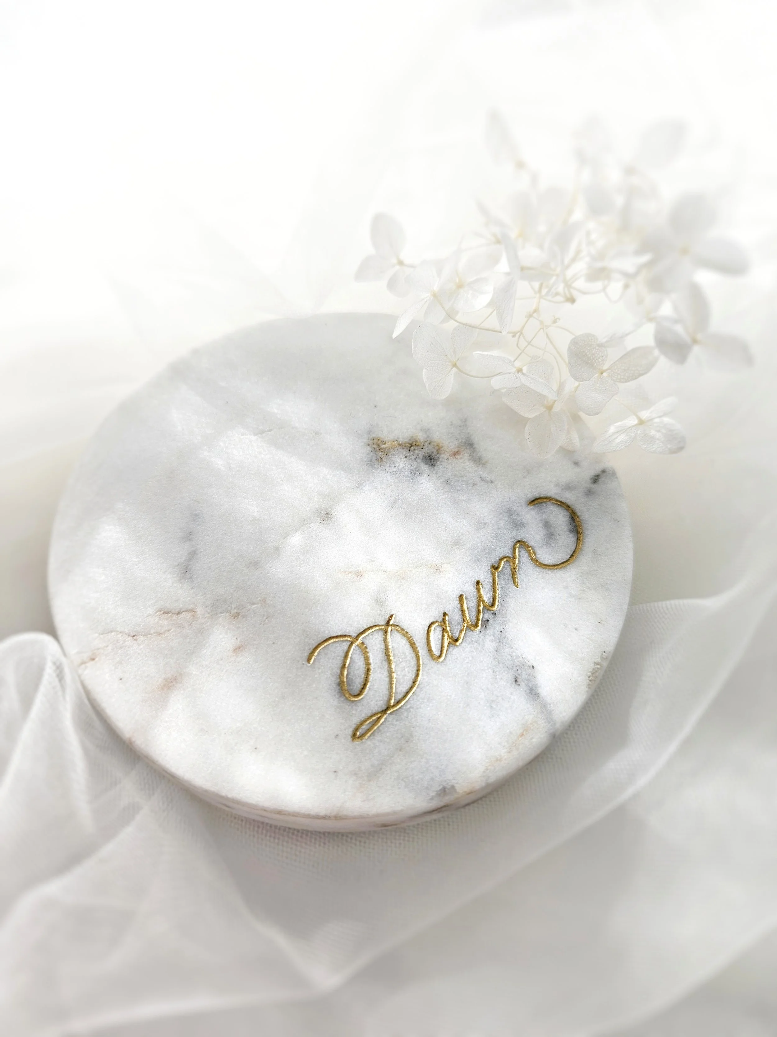 A white marble round object with the word 'Dream' in gold script, accented with a small gold crescent moon. Decorative white flowers and sheer fabric are also in the image.