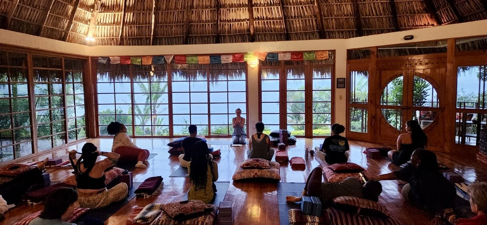 Ashley Smaldino teaching a group yoga class in a yoga studio with large windows showing palm trees and an ocean view.