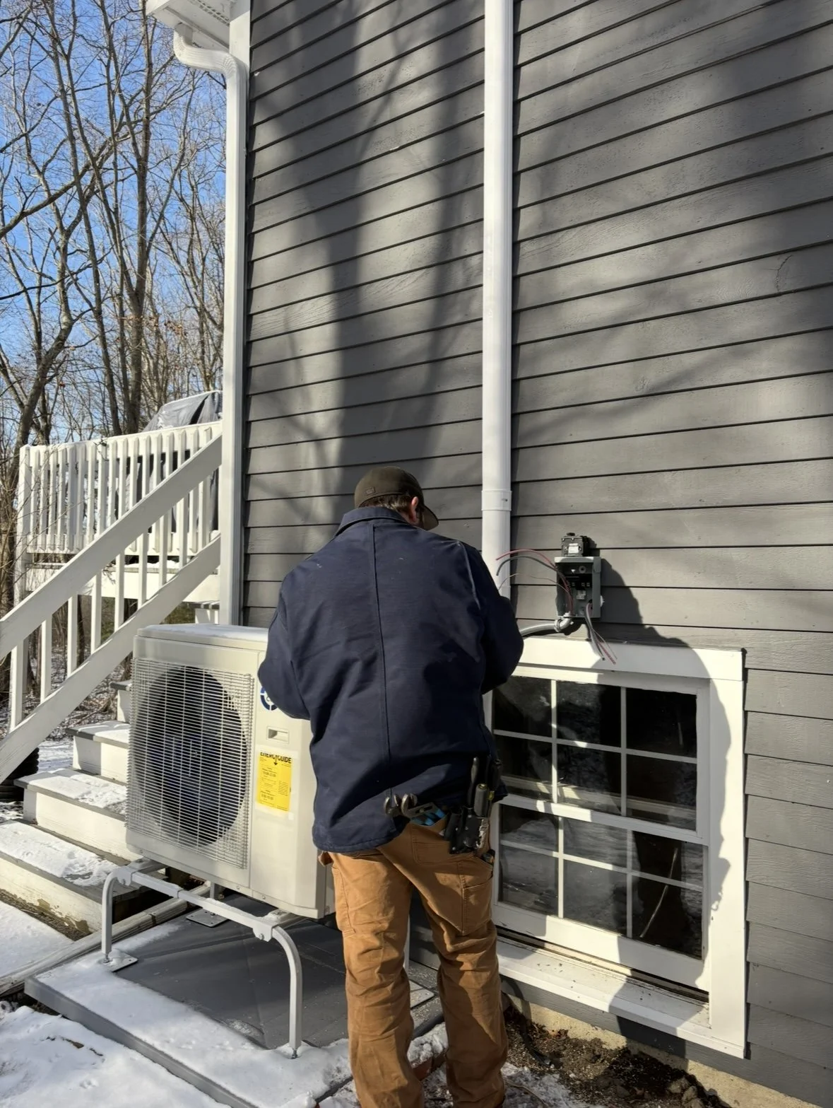 Technician working on outdoor electrical wiring near a window at a house in winter, with an air conditioning unit nearby.
