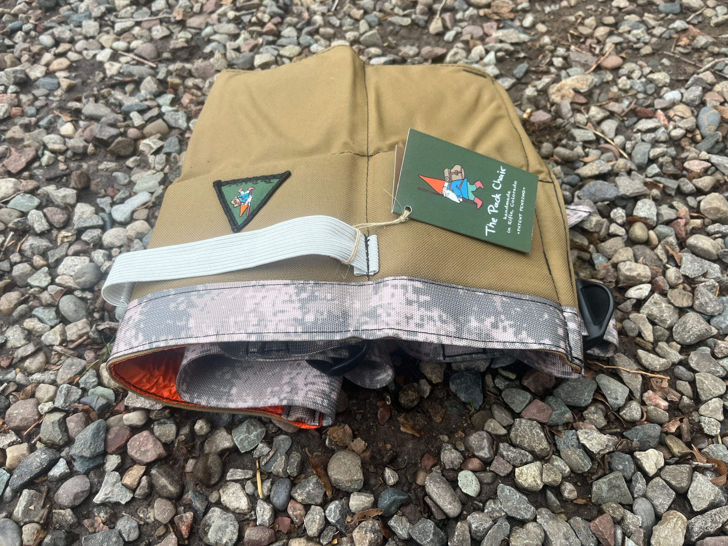 A tan Pack Chair with a white strap and a gray camouflage pattern near the bottom, lying on gravel ground. The Pack Chair has a green triangular patch and a green tag attached, which features a graphic of a gnome and the text 'The Pack Chair'.