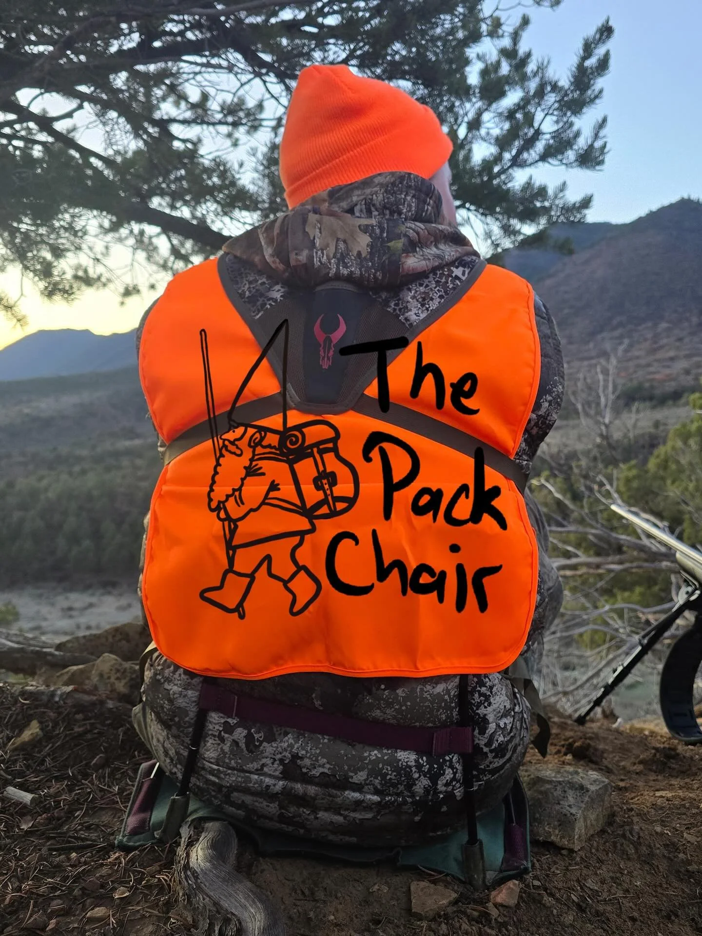 A person in camouflage clothing and an orange beanie and vest, sitting outdoors next to mountains and trees, with a drawing of a hiker carrying a pack, and text that reads "The Pack Chair" on the vest.