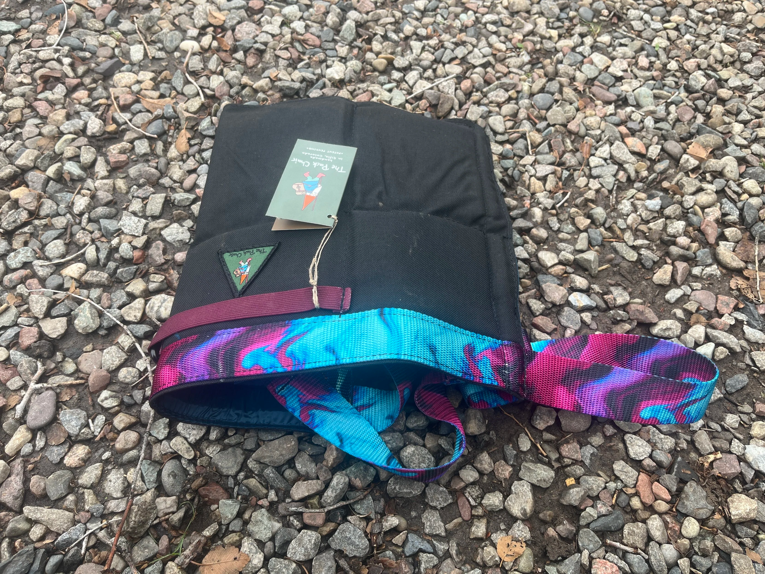 A black Pack Chair with colorful, patterned straps lying on a gravel surface.