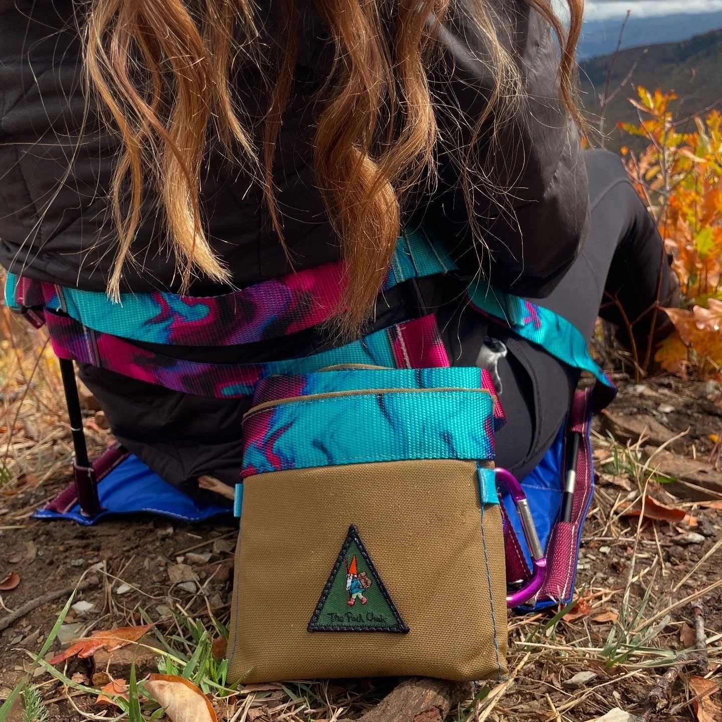A woman sitting on rocks by a river with lush greenery, trees, and a small waterfall in the background.
