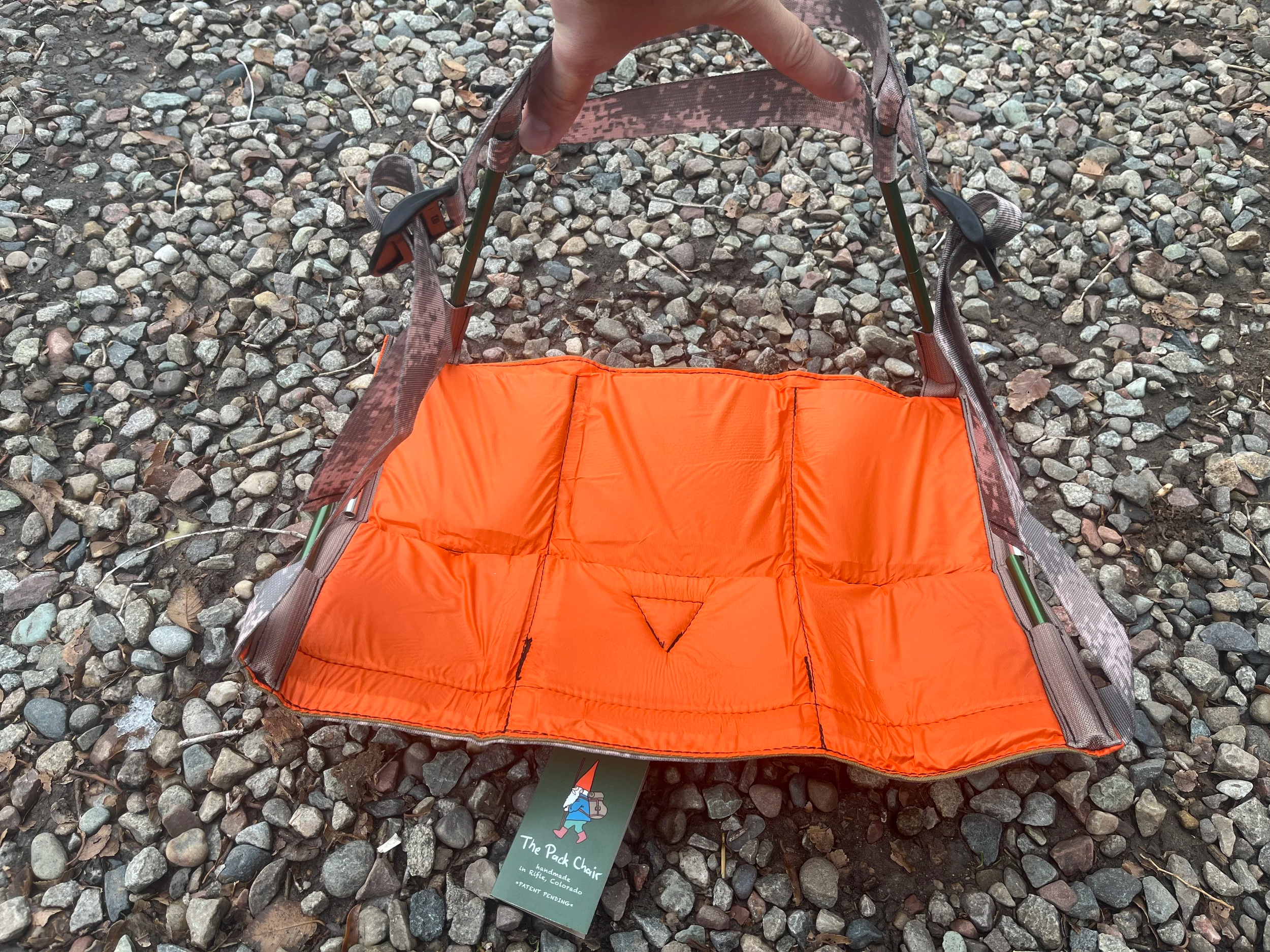 Orange padded foldable camping chair on rocky ground with a tag that reads "The Pack Chair".
