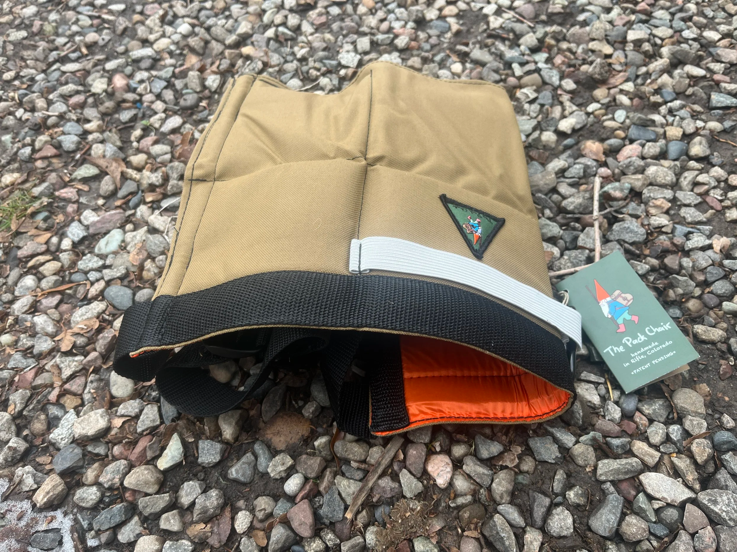 A tan and black Pack Chair with a white strap and orange interior lying on a gravel surface.