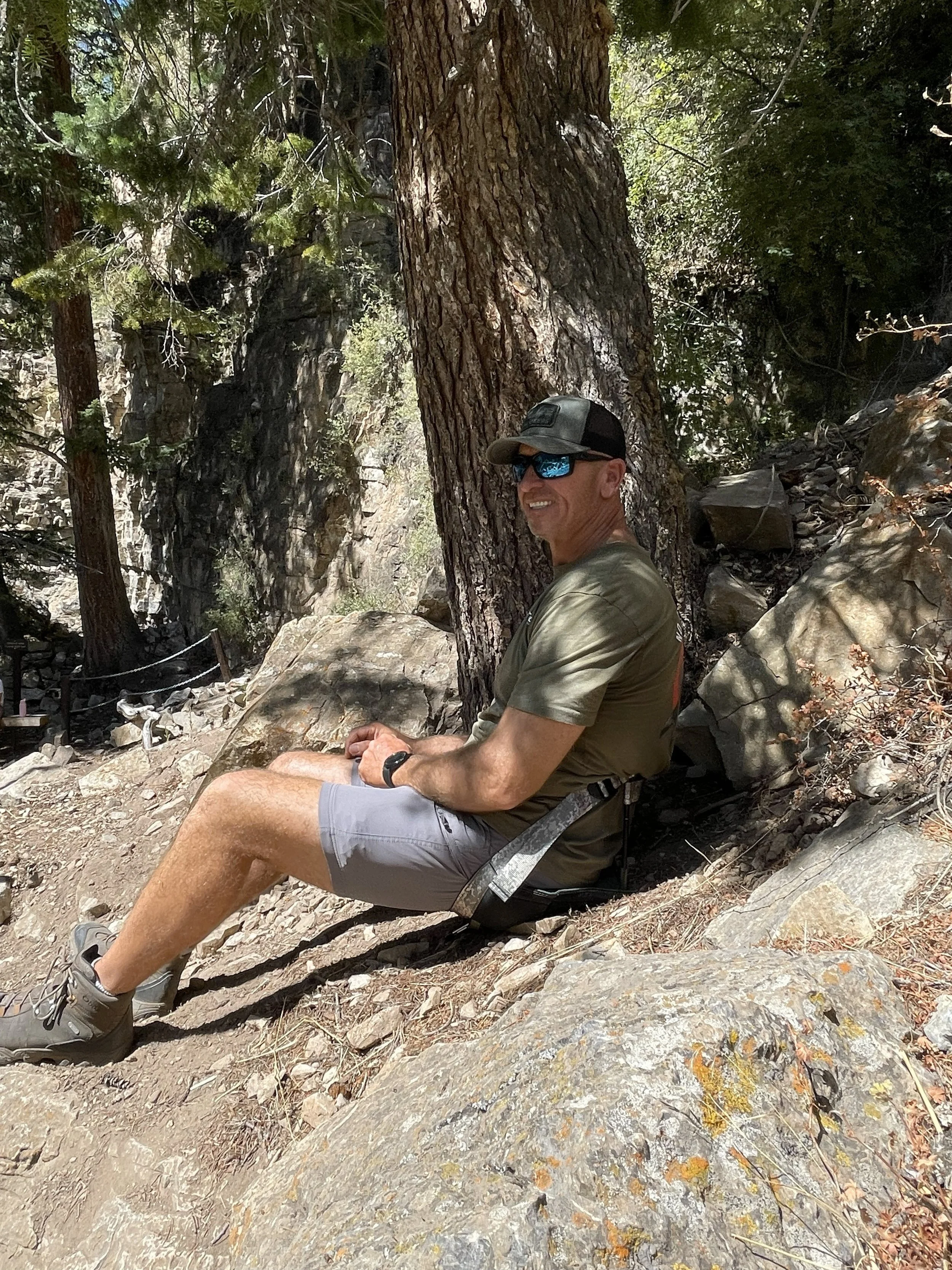 A man wearing sunglasses, a baseball cap, a green t-shirt, gray shorts, and hiking boots sits on a rock, smiling at the camera, in a forested area with large trees and rocks.
