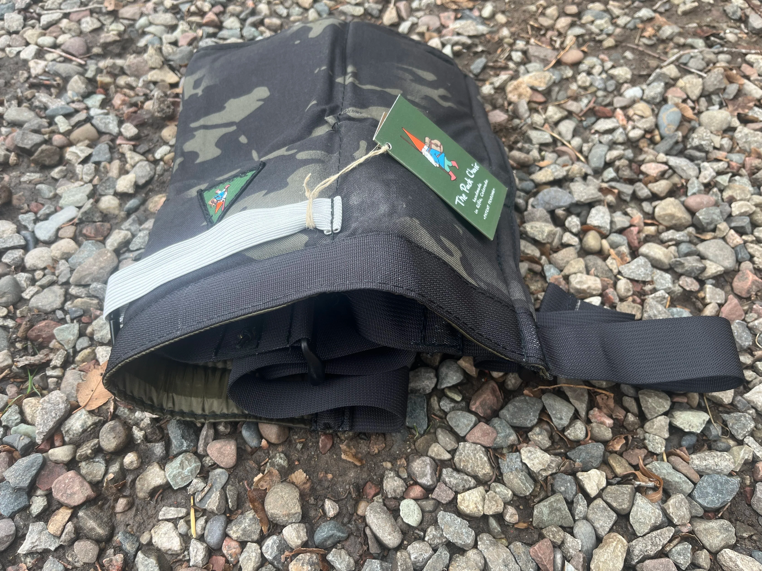 A camouflage-patterned Pack Chair with a black strap, lying on a bed of small rocks and leaves.