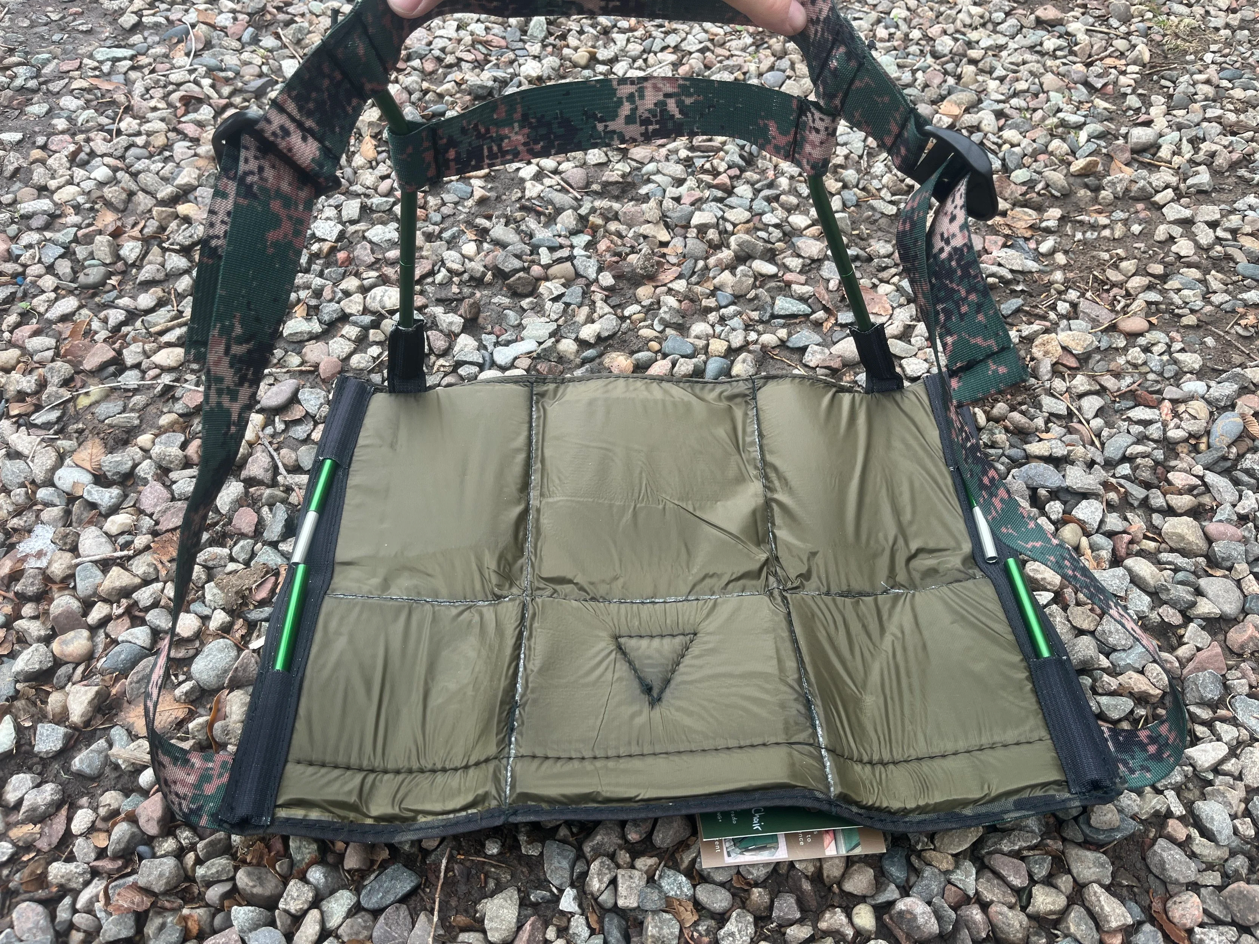 A portable, foldable seat with a padded, olive green cushion and adjustable straps, placed on a gravel surface outdoors.