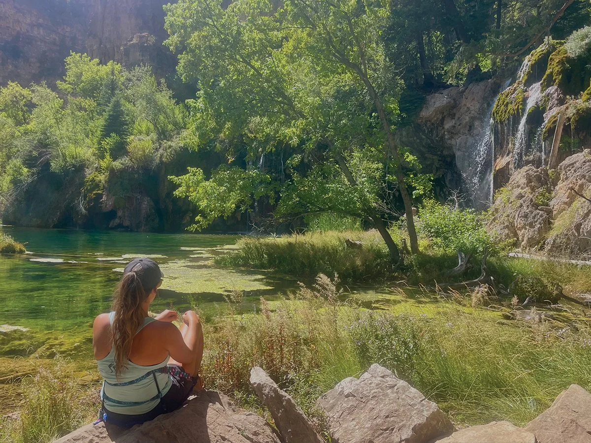 A woman sitting on rocks by a river with lush greenery, trees, and a small waterfall in the background.