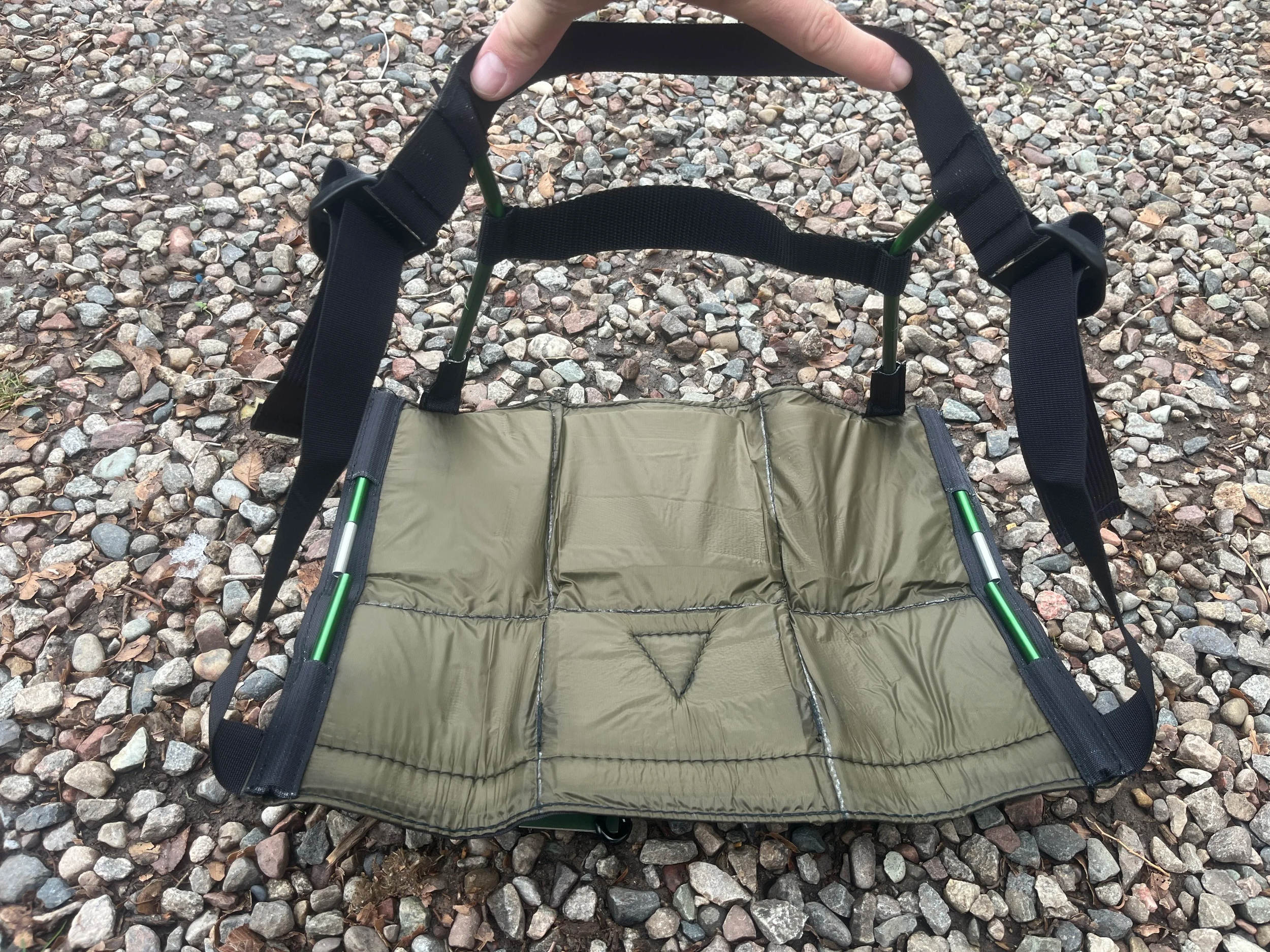 A portable folding seat with a tan cushion and black straps, positioned on a gravel surface.