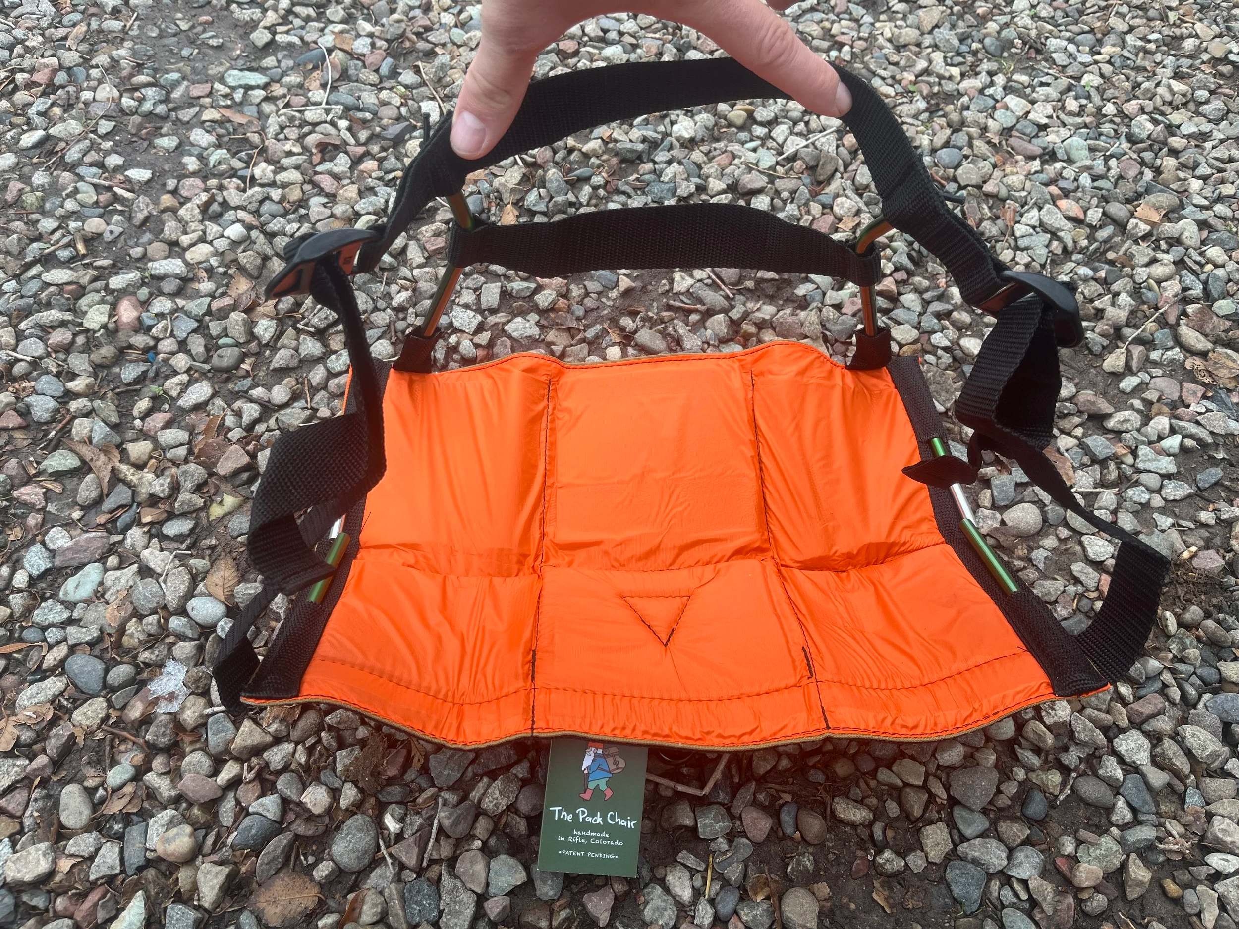 Hand holding an orange pack chair with black straps on a gravel ground.