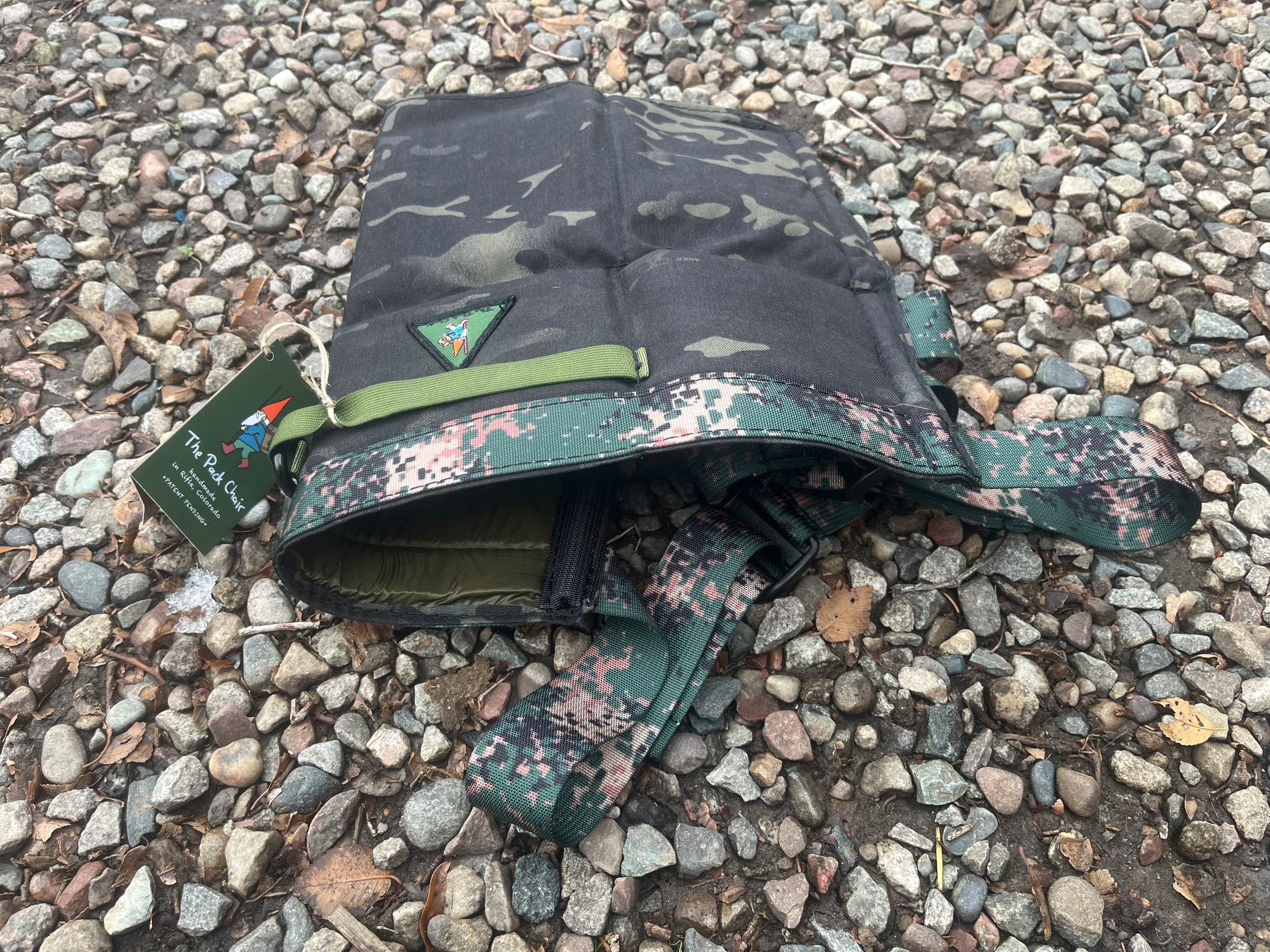 A black camouflage-patterned Pack Chair lying on a gravel ground with a green tag attached.