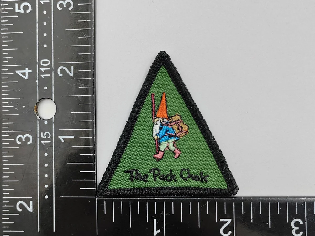A green triangular embroidered patch with a black border, featuring a gnome carrying a backpack and wearing a bright orange hat, with the text "The Ped Chat" embroidered at the bottom.