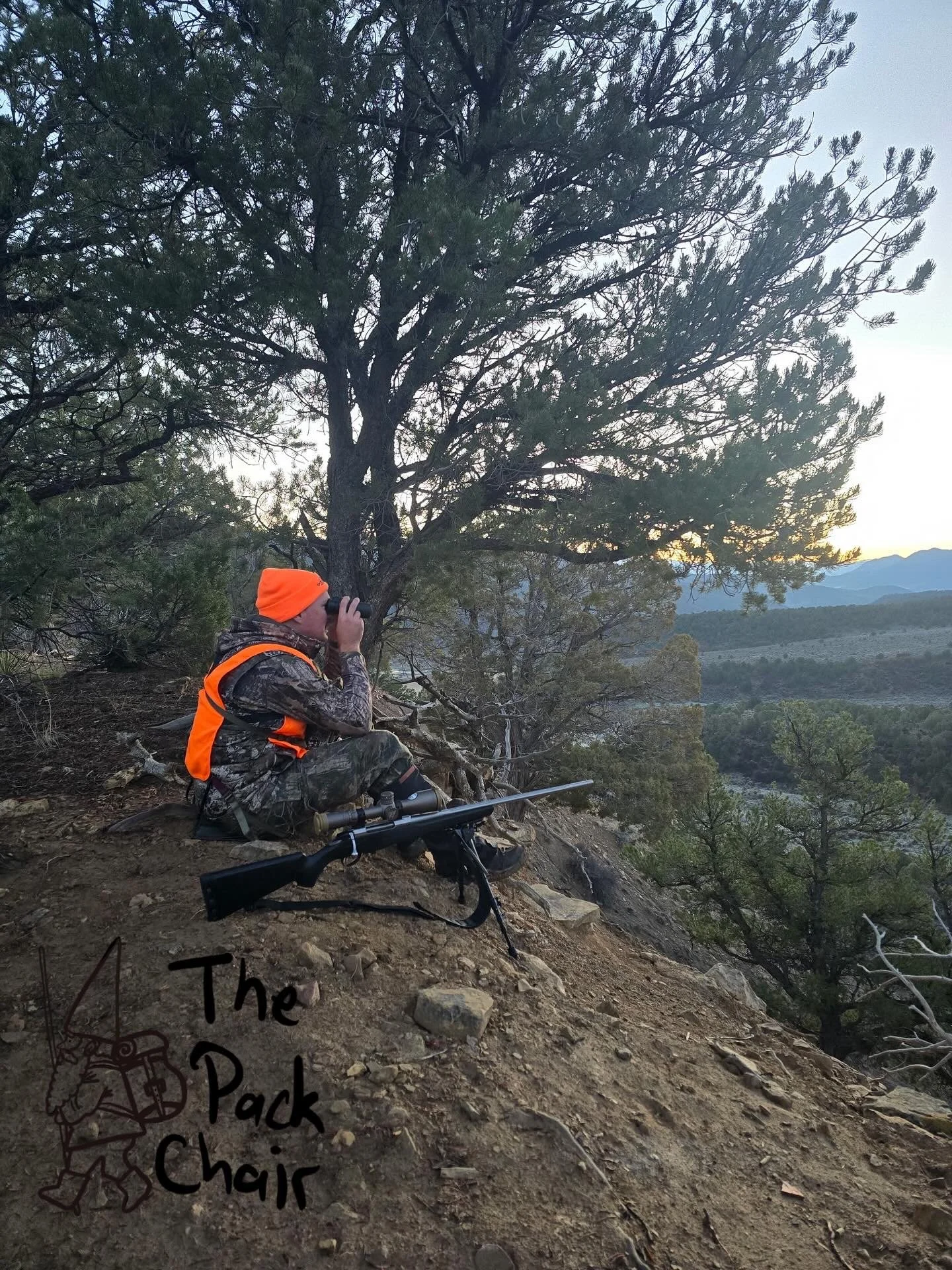 A person dressed in camouflage and an orange jacket and hat, sitting on a dirt slope during sunset, looking through binoculars at the landscape, with a camera and a rifle nearby, and a logo for 'The Pack Chair' in the bottom left corner.