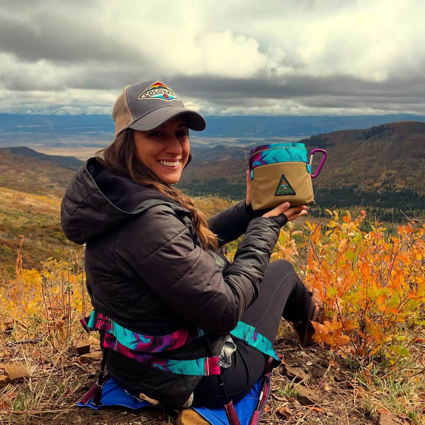 A woman sits on a hillside with orange autumn foliage, holding a colorful insulated cup and smiling at the camera. The background features distant mountains and a cloudy sky.