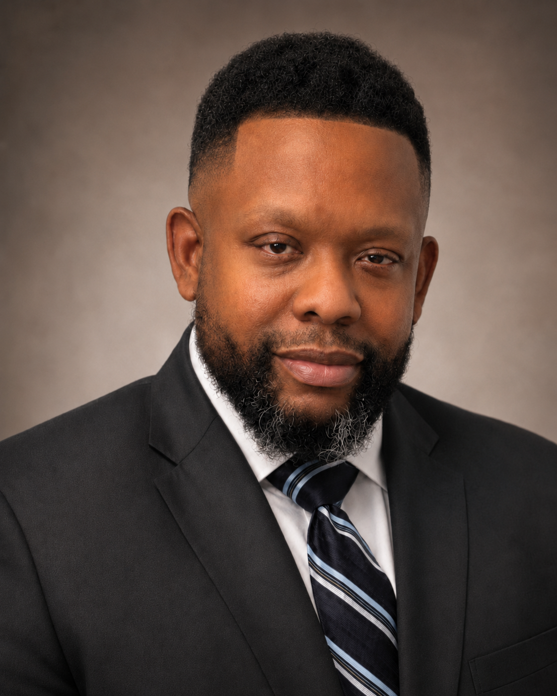 A professional headshot of a Black man with a beard, wearing a black suit, white shirt, and a striped tie, against a neutral background.