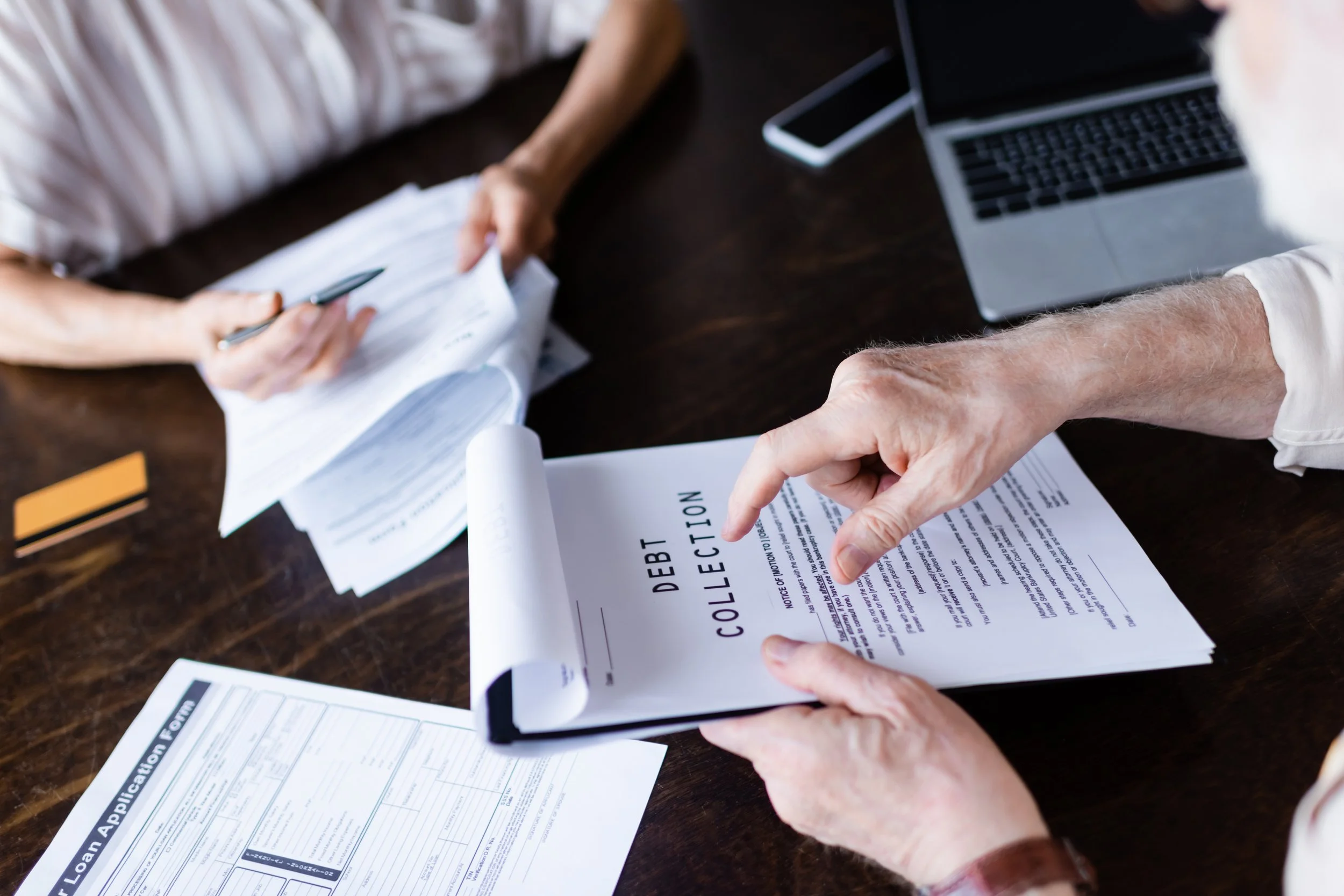 Two people reviewing debt collection and loan application documents on a dark wooden table, with a laptop, a smartphone, and a yellow card present.