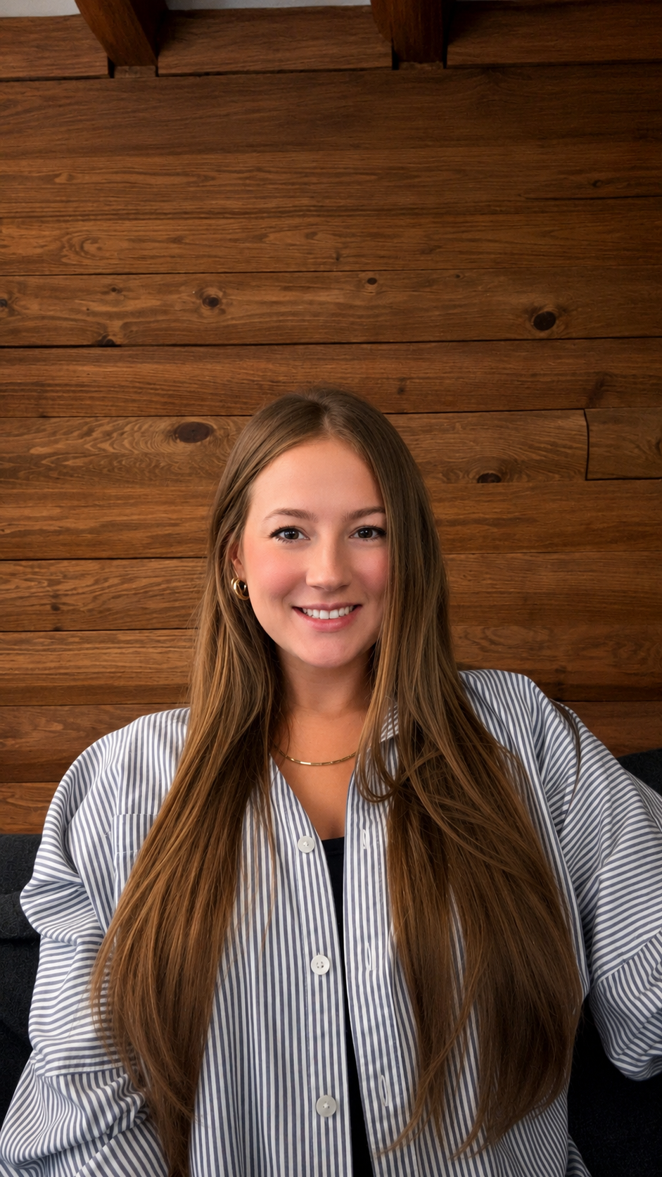 A young woman with long brown hair, wearing a striped button-up shirt, gold hoop earrings, and a gold necklace, smiling in front of a wooden wall.