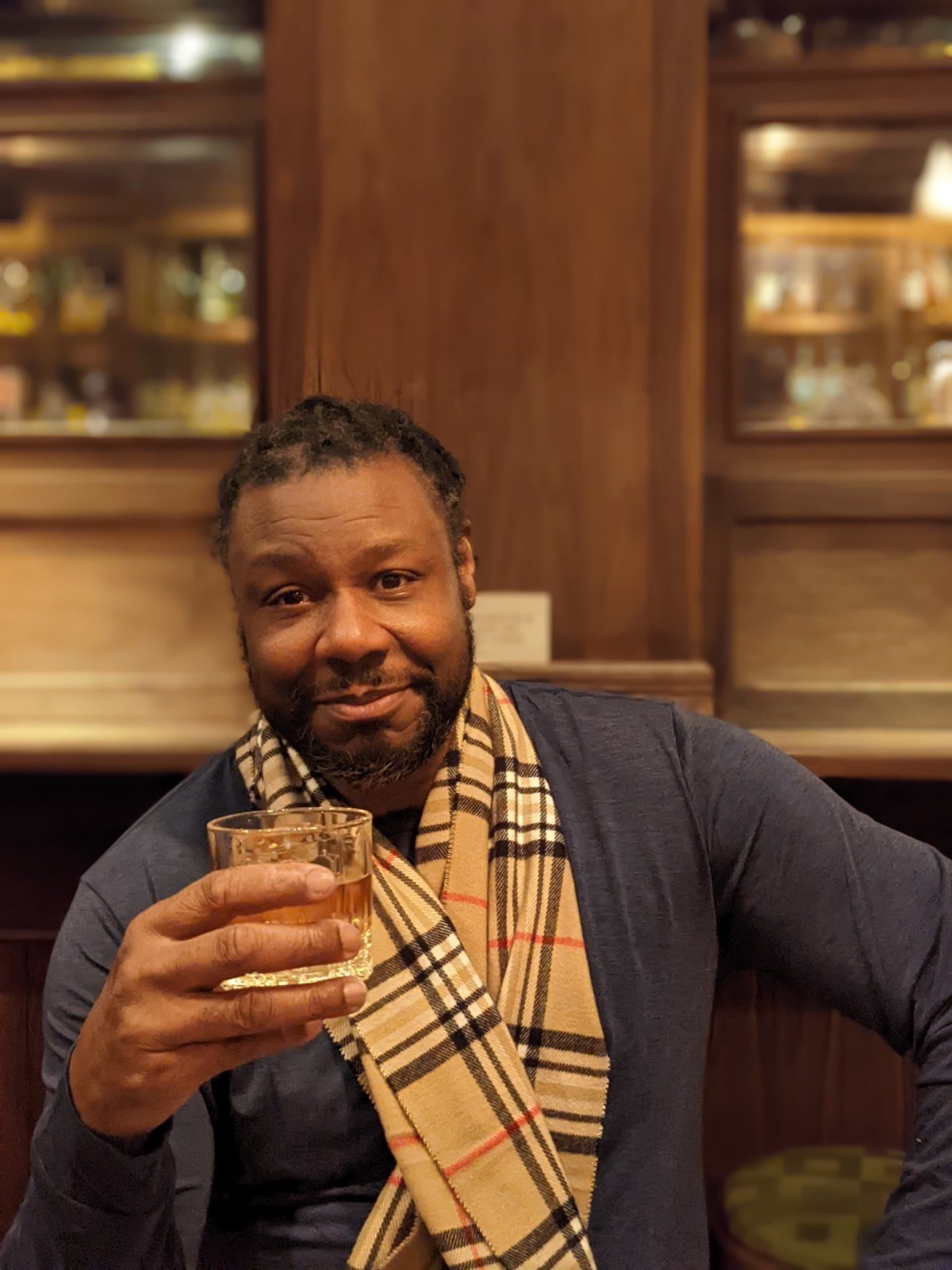 A Vincent Keele toast to all my collectors, patrons and art lovers. with short, Wearing a dark-colored shirt and a beige Burberry scarf, smiling and holding a glass of whiskey in a warmly lit, wood-paneled in a Bronx restaurant.