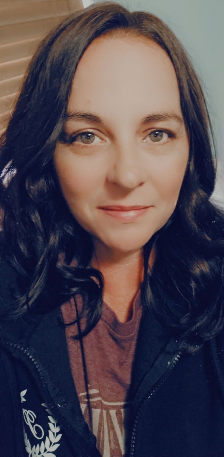 Close-up portrait of a woman with dark wavy hair, light skin, and blue eyes, smiling slightly, wearing a dark zip-up jacket and a maroon shirt.