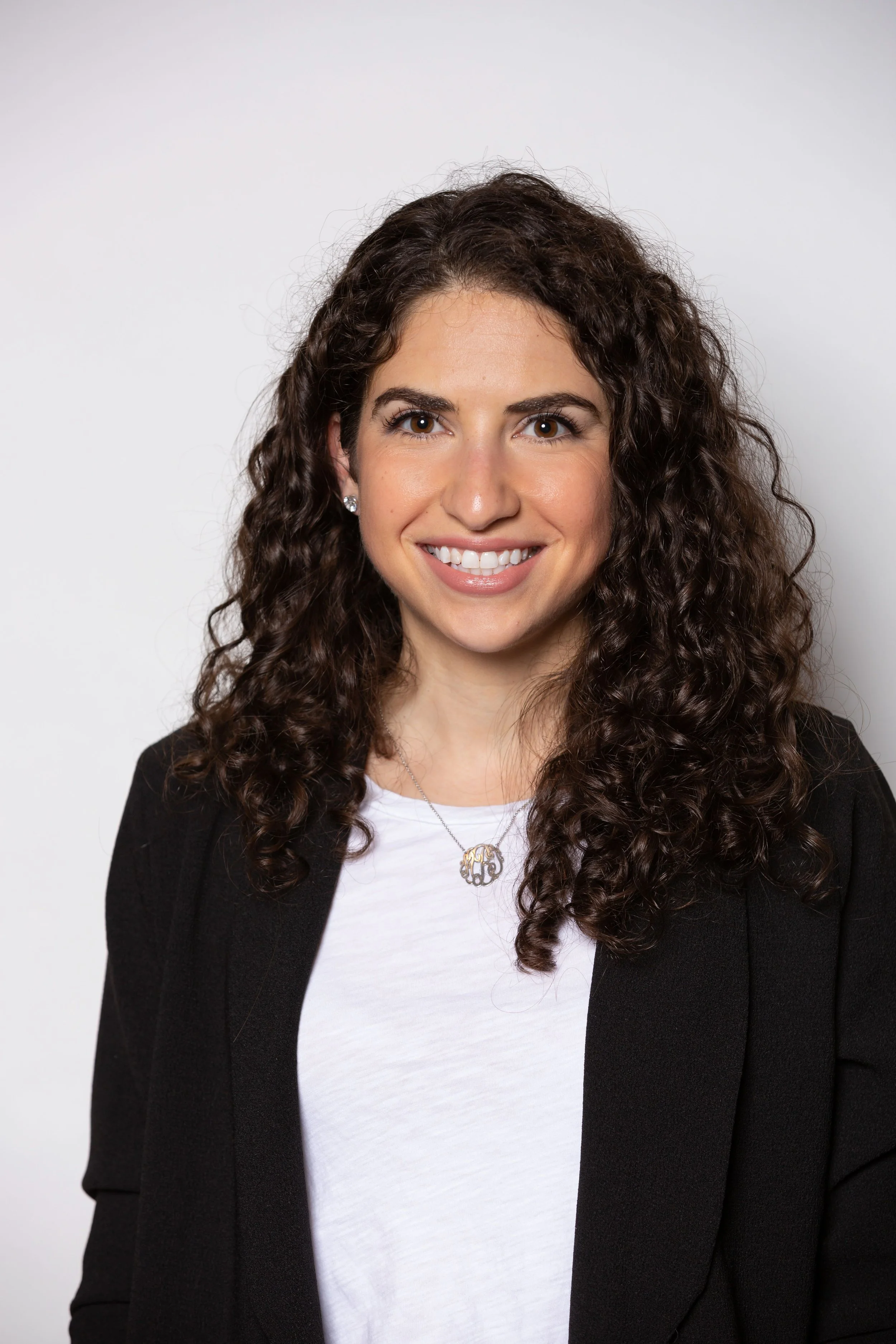 Image of a woman with light skin and dark brown curly hair, facing camera and smiling. Wearing a white shirt under a black blazer.